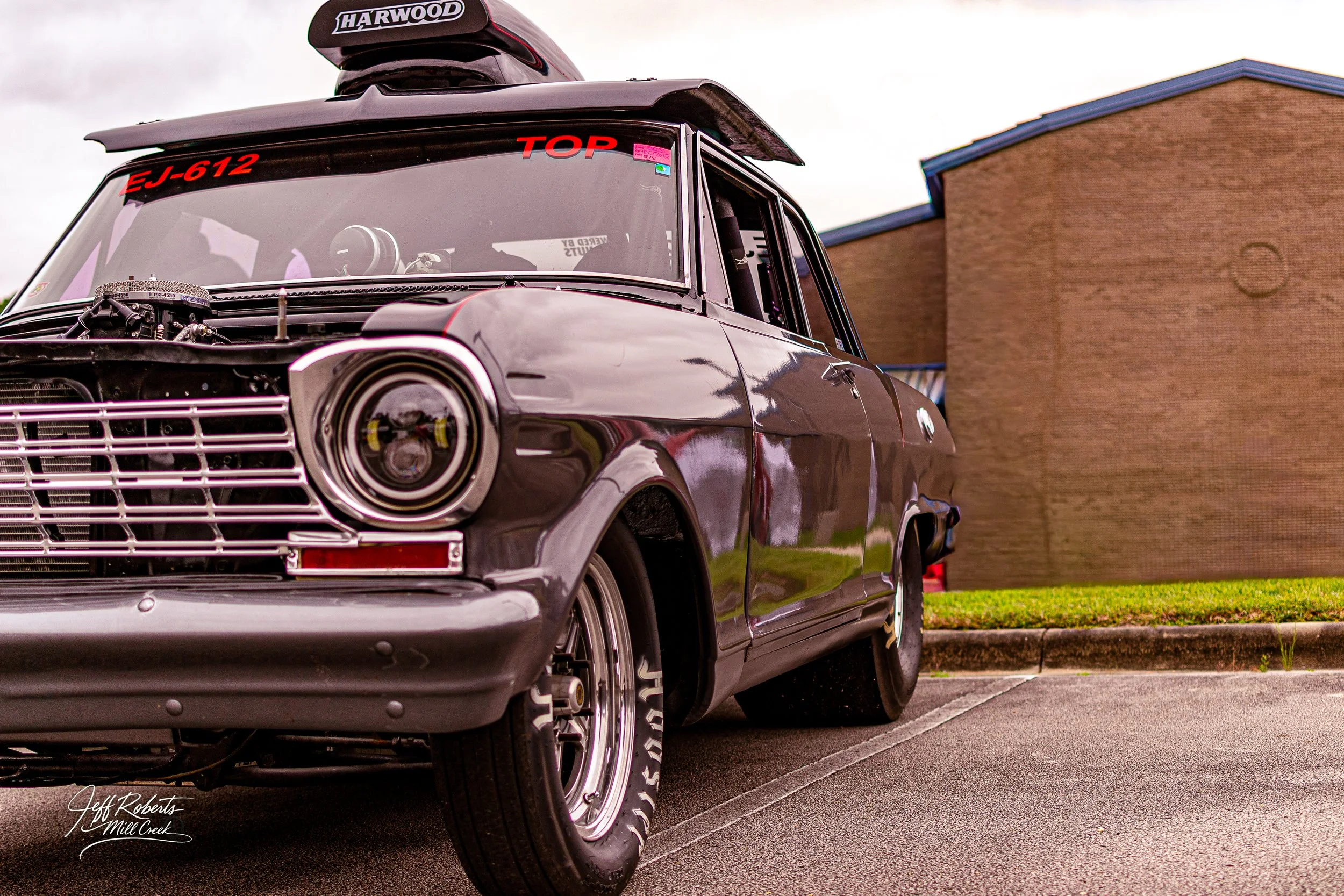 A vintage black muscle car with a custom front grille and a large rear spoiler, parked on a parking lot in front of a brick building, featuring racing decals and modifications.
