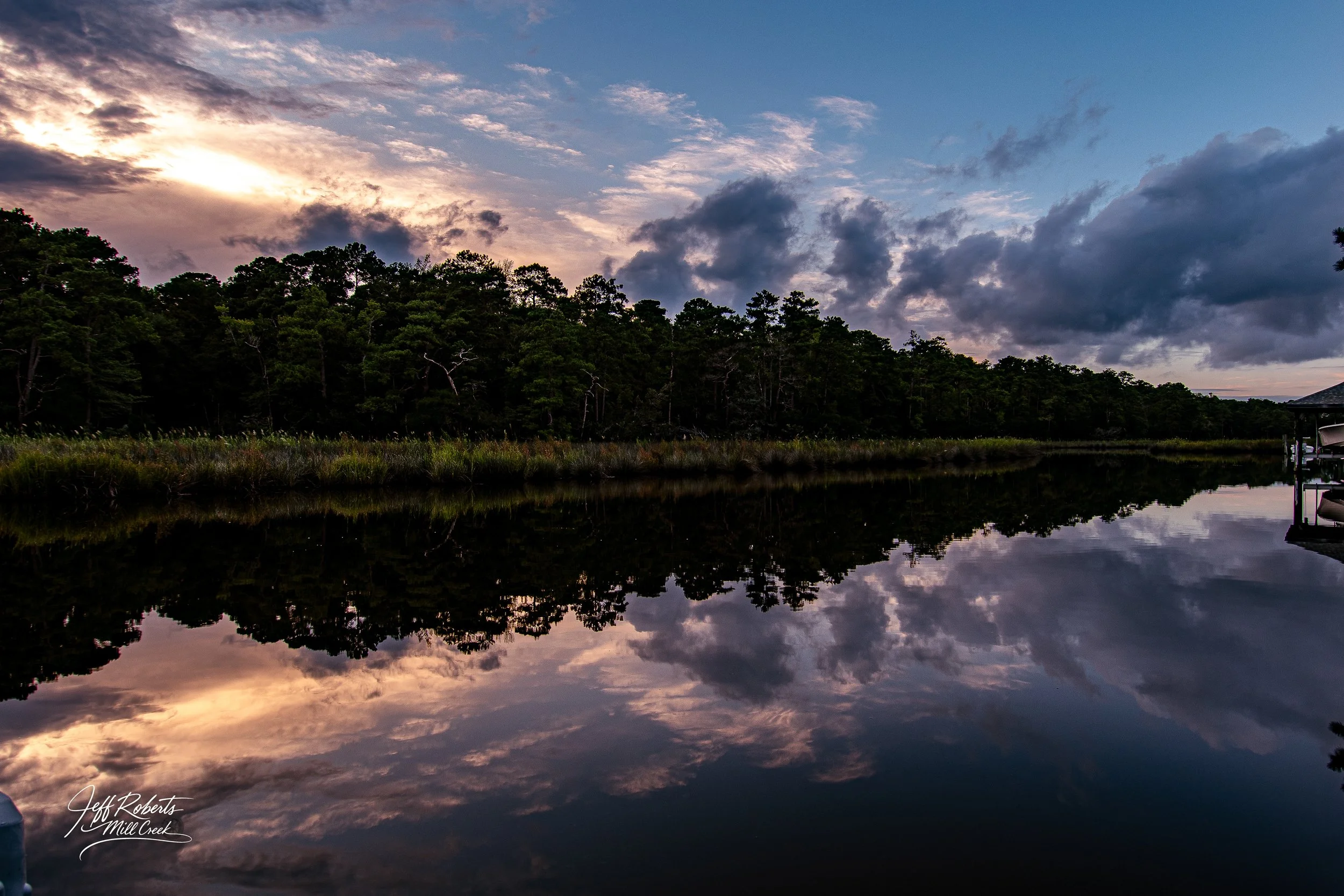Sunset over a calm river with cloud reflections, trees on the far bank, and a small dock on the right, signature in lower left corner.