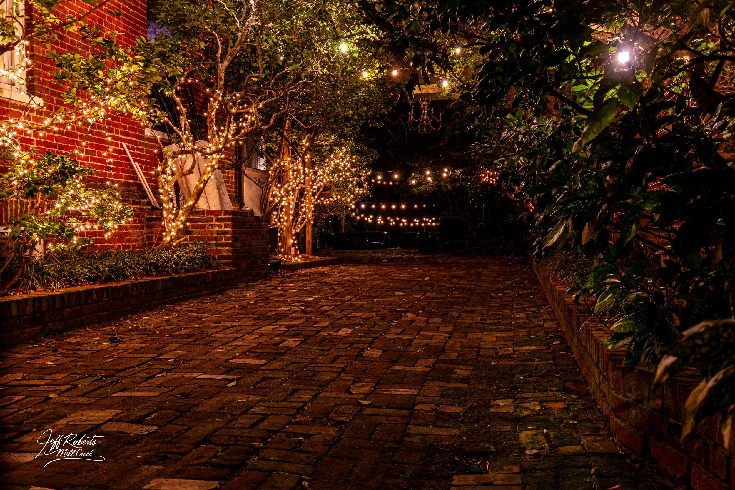 A cobblestone path lined with trees wrapped in string lights and a brick building side covered in lights, creating a cozy, festive atmosphere at night.