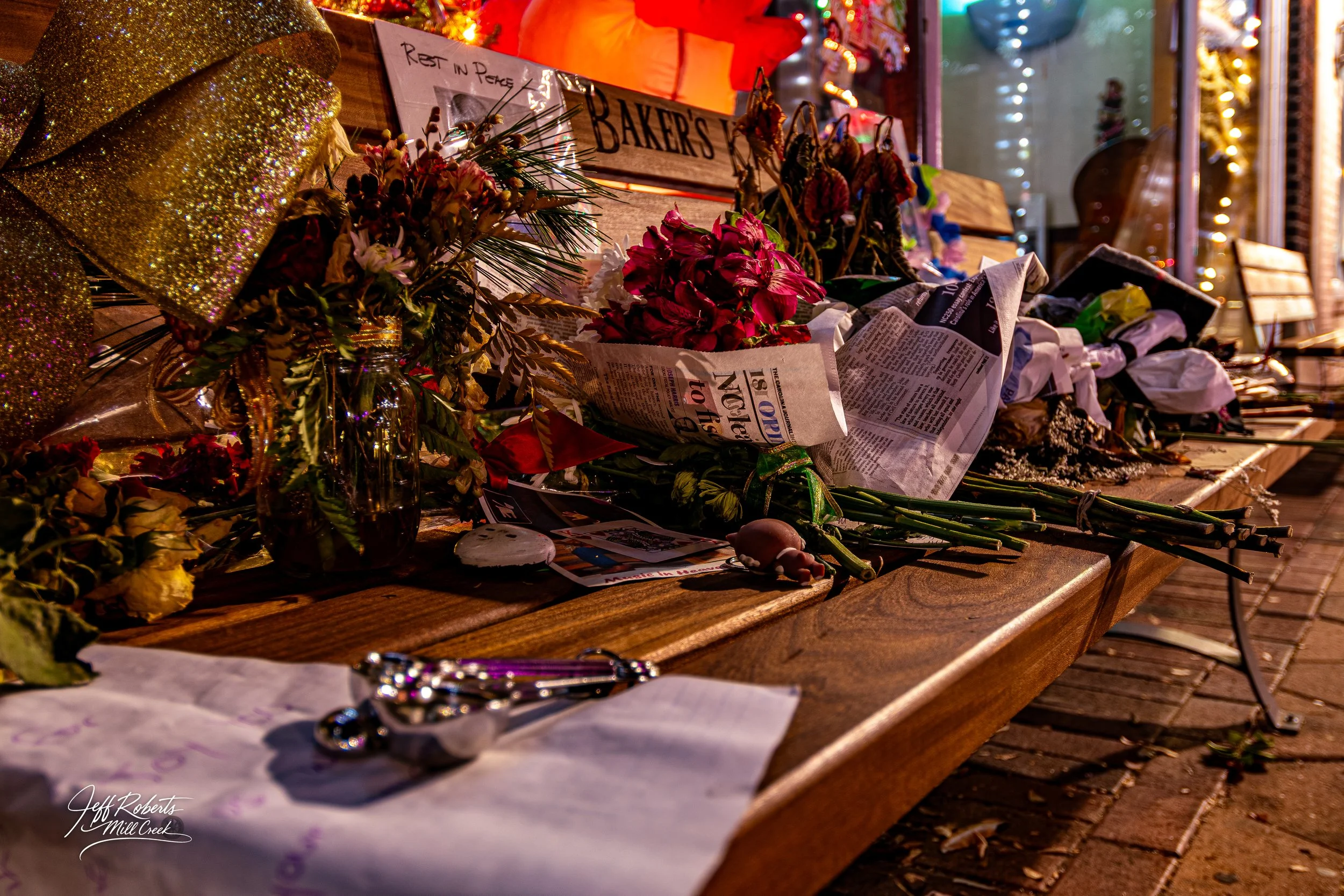 A memorial display on a wooden bench with flowers, handwritten notes, newspapers, and various items. There are Christmas lights and a decorated setting in the background, indicating a somber tribute scene.