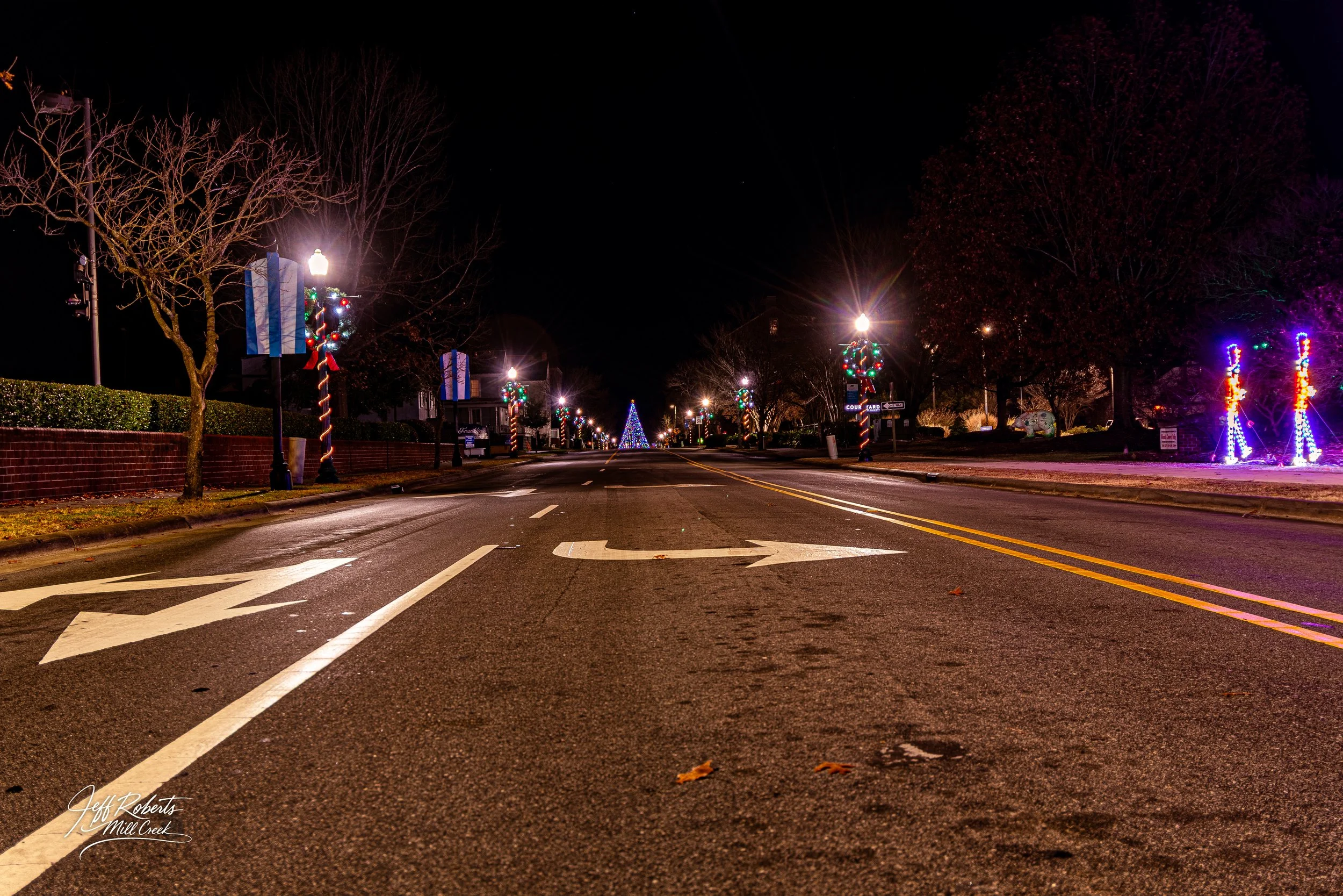 Empty street decorated with holiday lights, lit trees, and a Christmas tree in the distance, nighttime view.