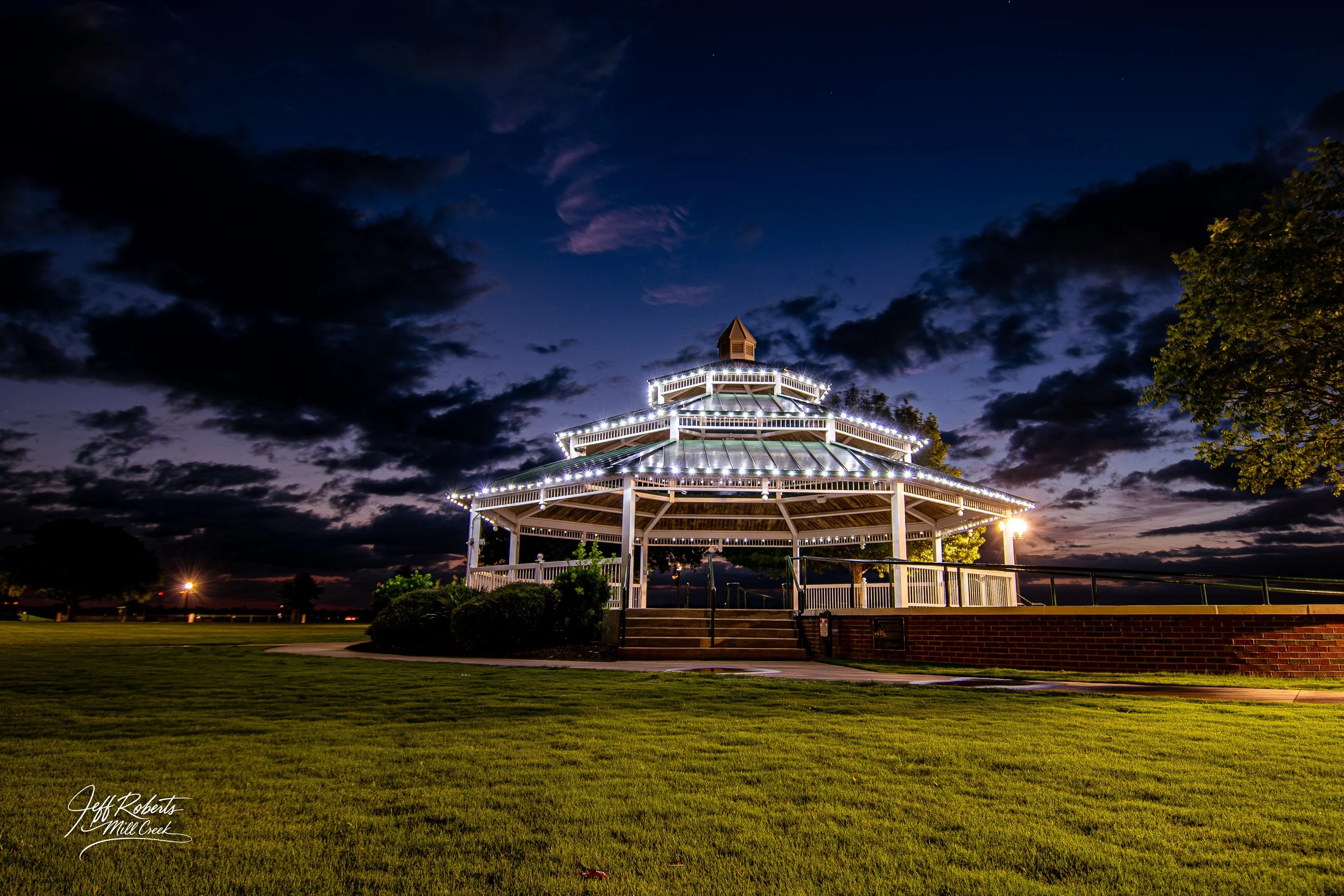 A decorated white gazebo with string lights in a park at dusk with a dark sky and clouds.