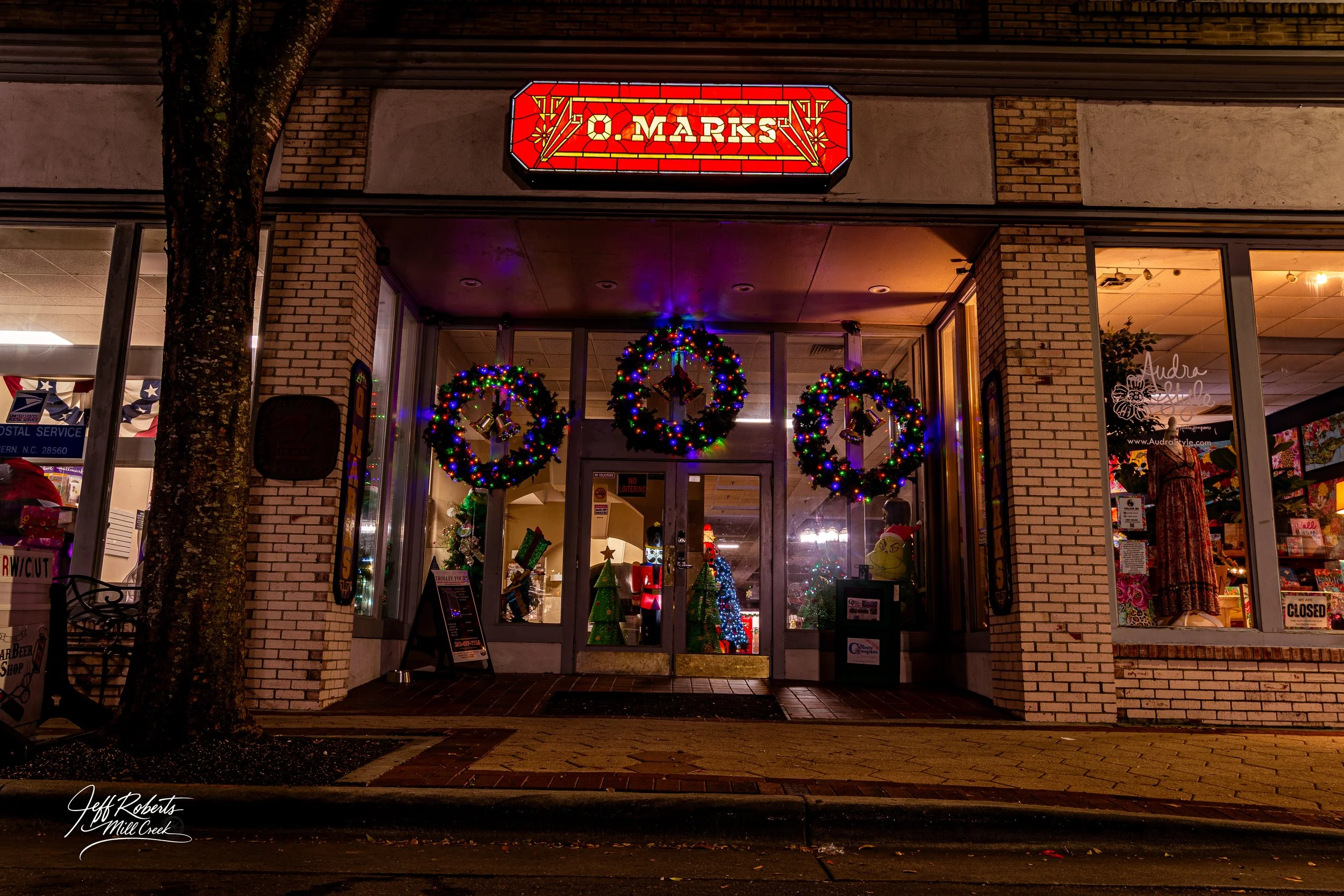 Night view of a storefront decorated with colorful Christmas wreaths and lights, with a sign reading 'O. Marks' above the entrance