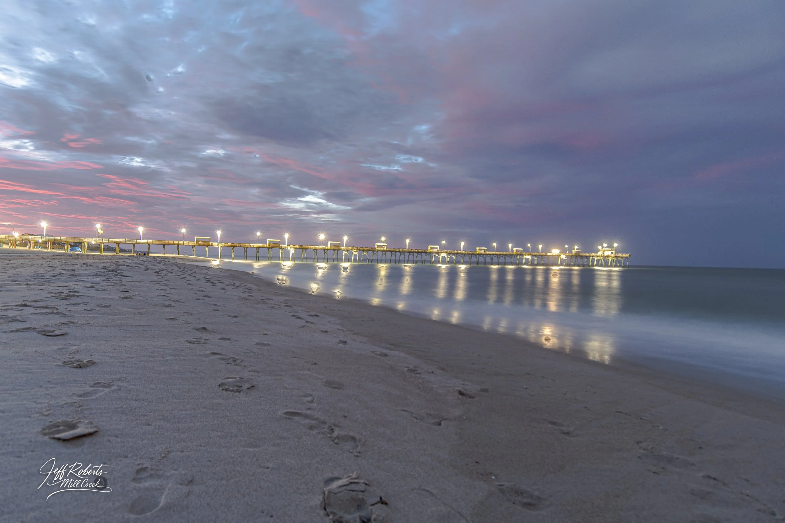 A beach with footprints on the sand during dusk, with a pier illuminated by lights extending into the ocean under a sky with pink, purple, and gray clouds.