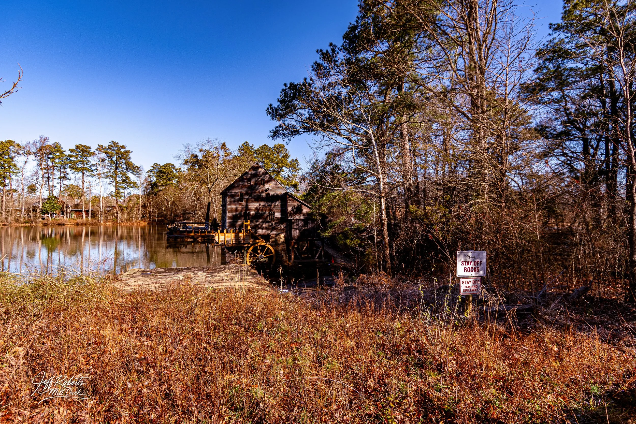 An old wooden mill house on a lake surrounded by trees and dry grass, with a sign that says 'Please stay off rocks' and 'Stay off dam and spillway'.