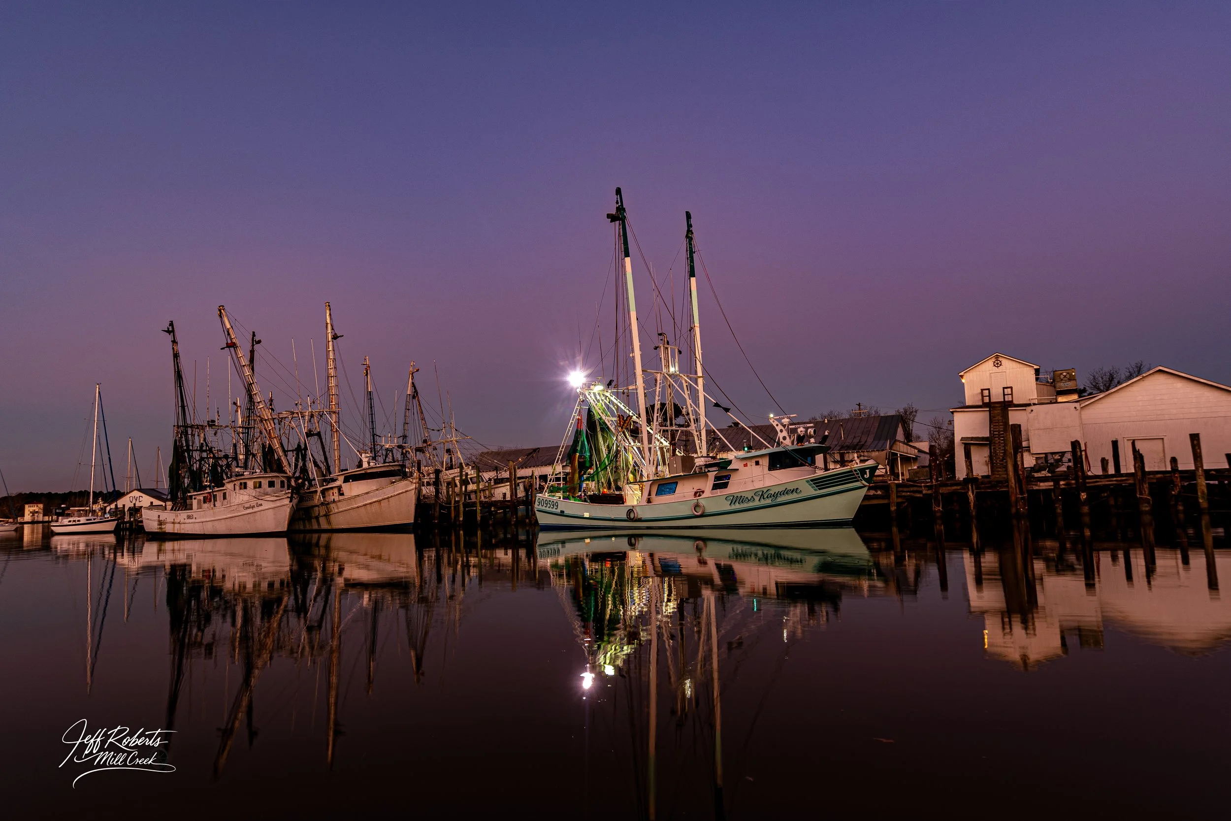 Boats docked at a marina during dusk with reflections in the calm water, and a building and houses in the background.