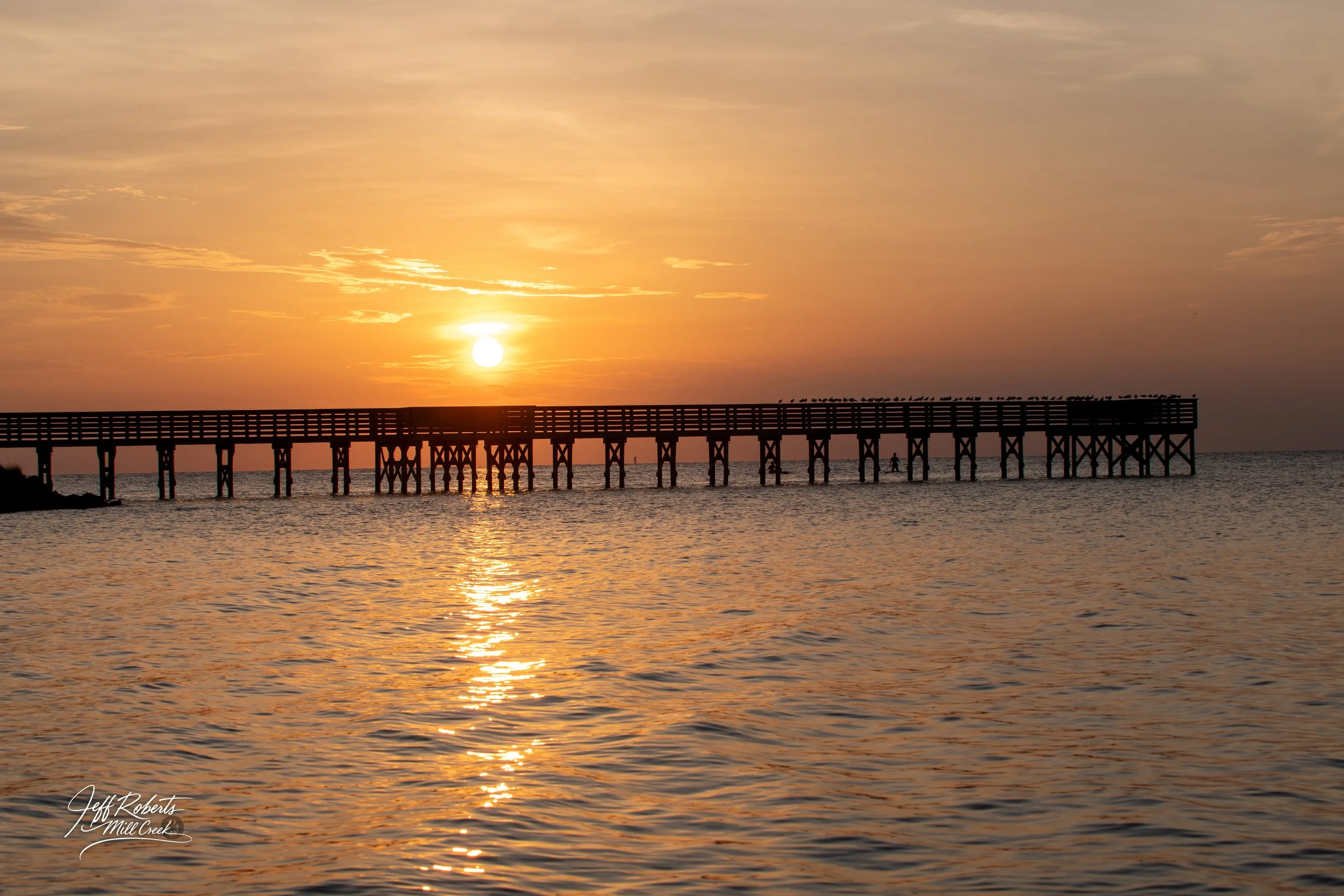 Sunset over the water with silhouettes of a long pier extending into the sea, some birds perched on the pier, and the sky filled with warm colors.
