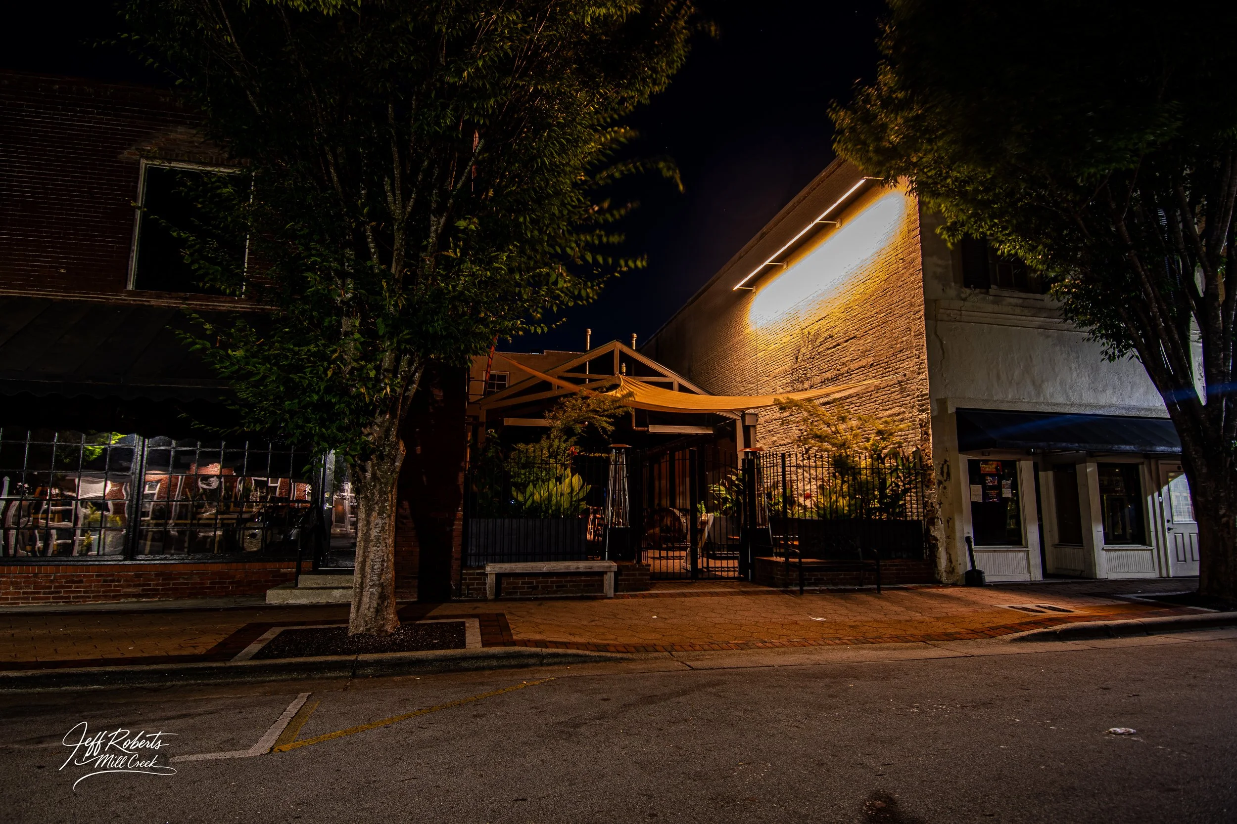 Night scene of a downtown street with a building illuminated by exterior lighting, trees in the foreground, and a parking lot on the left.