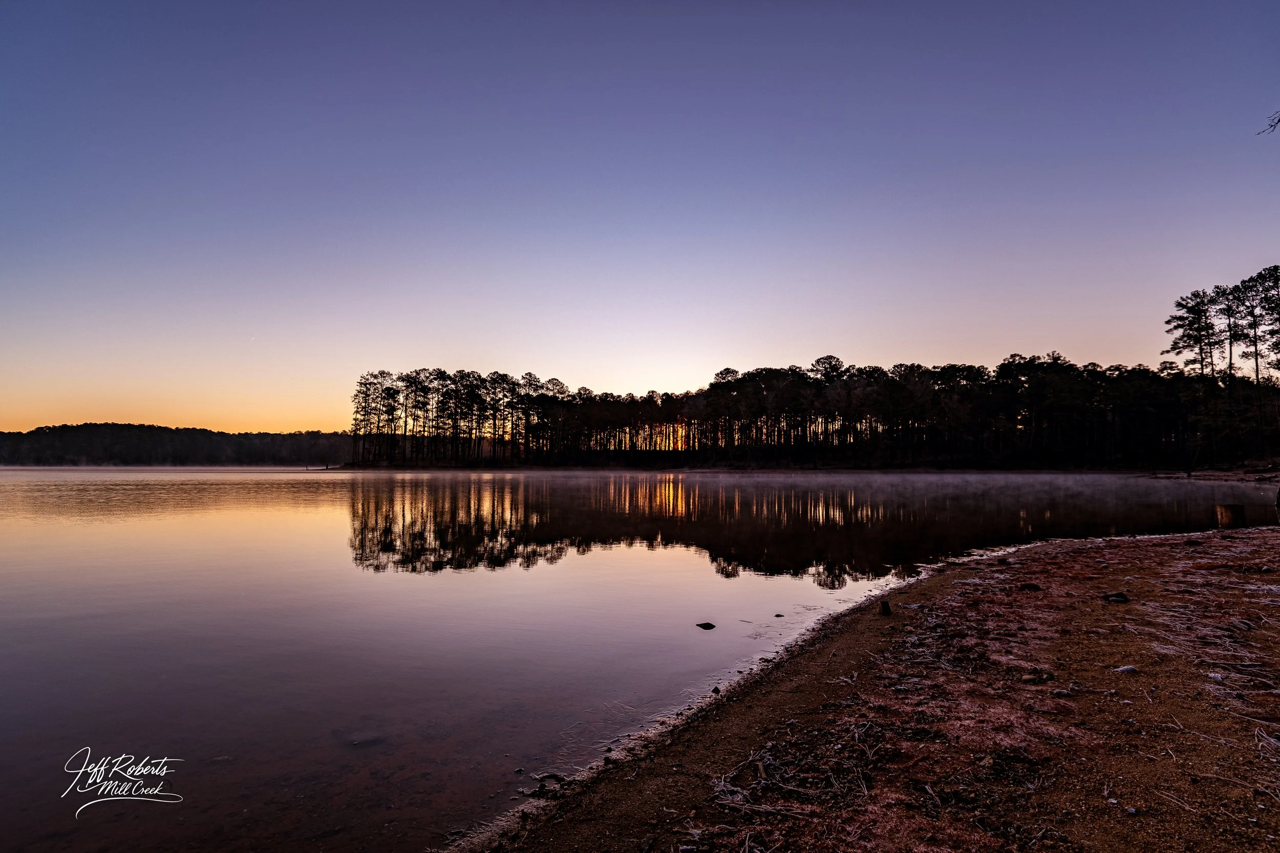 Sunset over a calm lake with a reflection of trees on the water, and a dirt shoreline in the foreground.