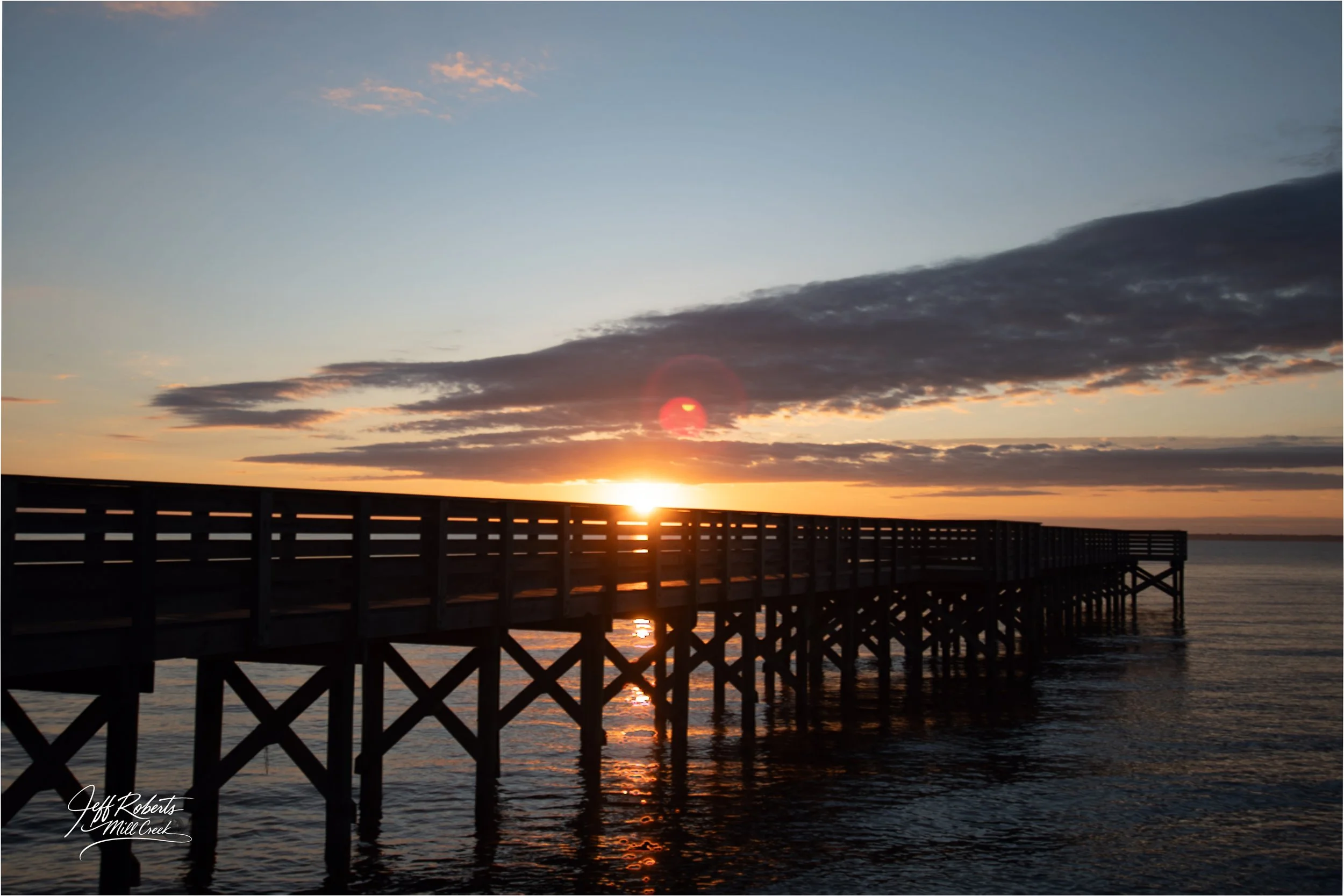 A wooden pier extending over water at sunset with the sun near the horizon and clouds in the sky.