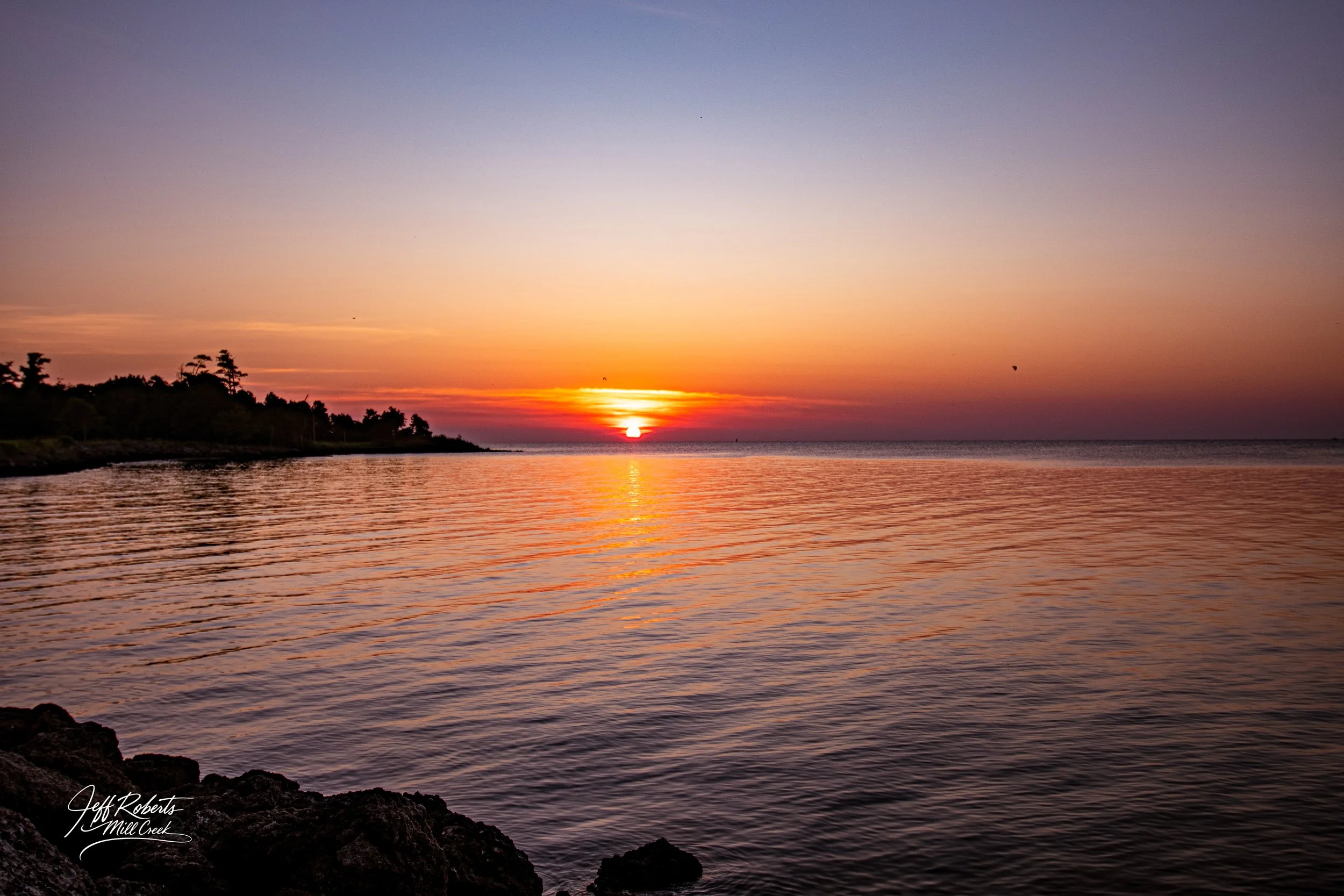 Sunset over a calm body of water with a rocky shoreline in the foreground and a tree-lined coast on the left.