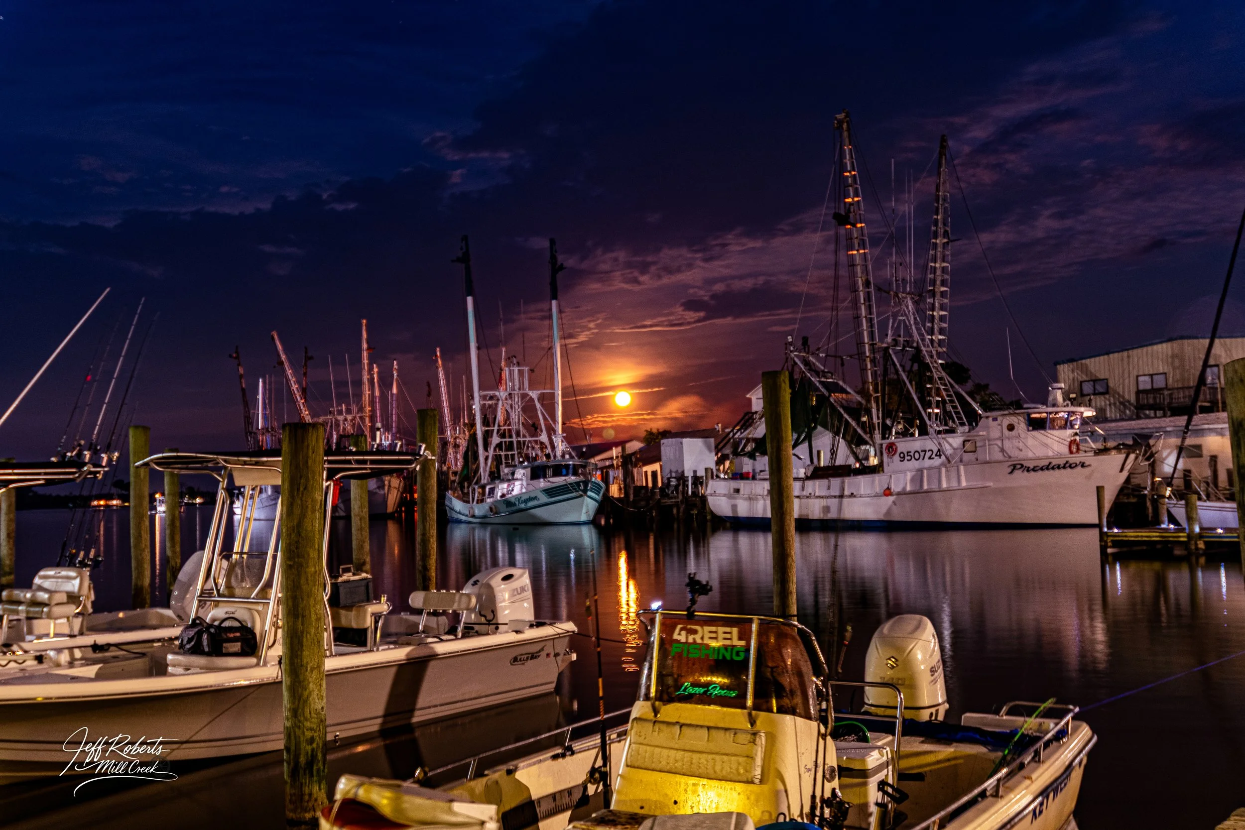Nighttime scene of boats docked at a marina with the moon in the sky and reflection on the water.