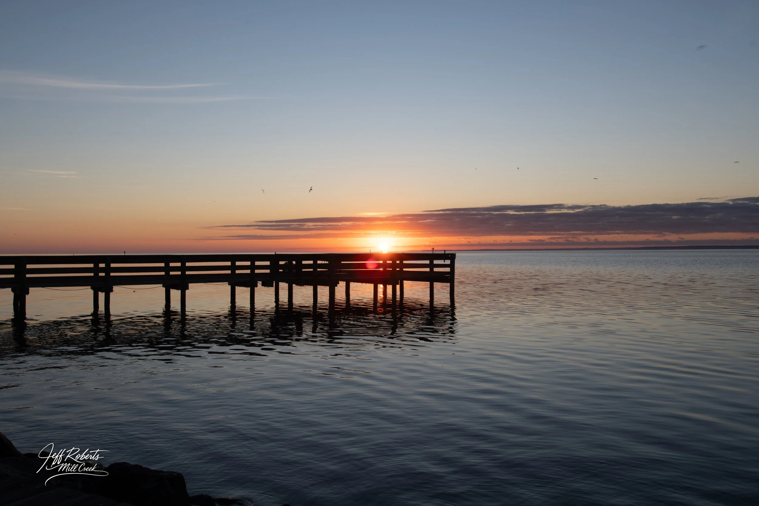 Sunset over a calm body of water with a wooden dock extending into the water.