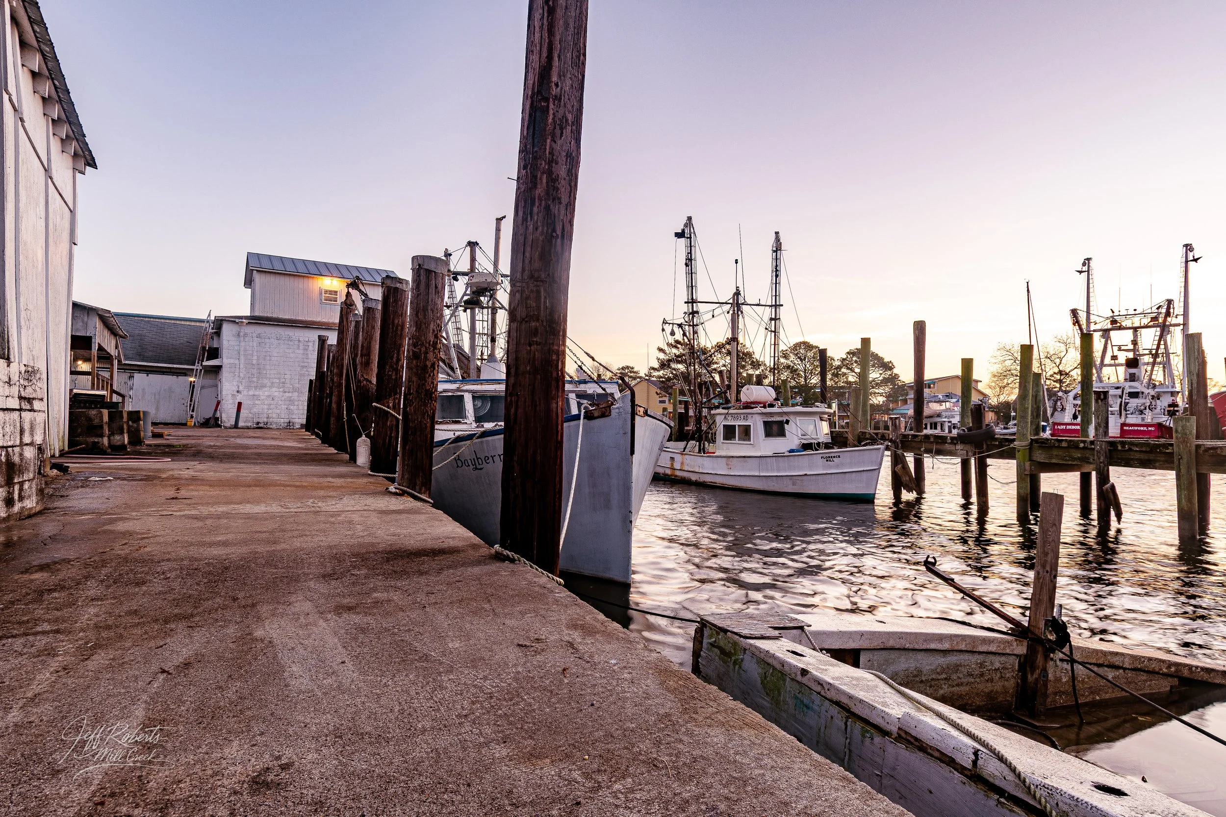A waterfront scene at sunset with boats docked along a wooden pier, some wooden posts, and small buildings on the left, calm water, and sky with soft colors.