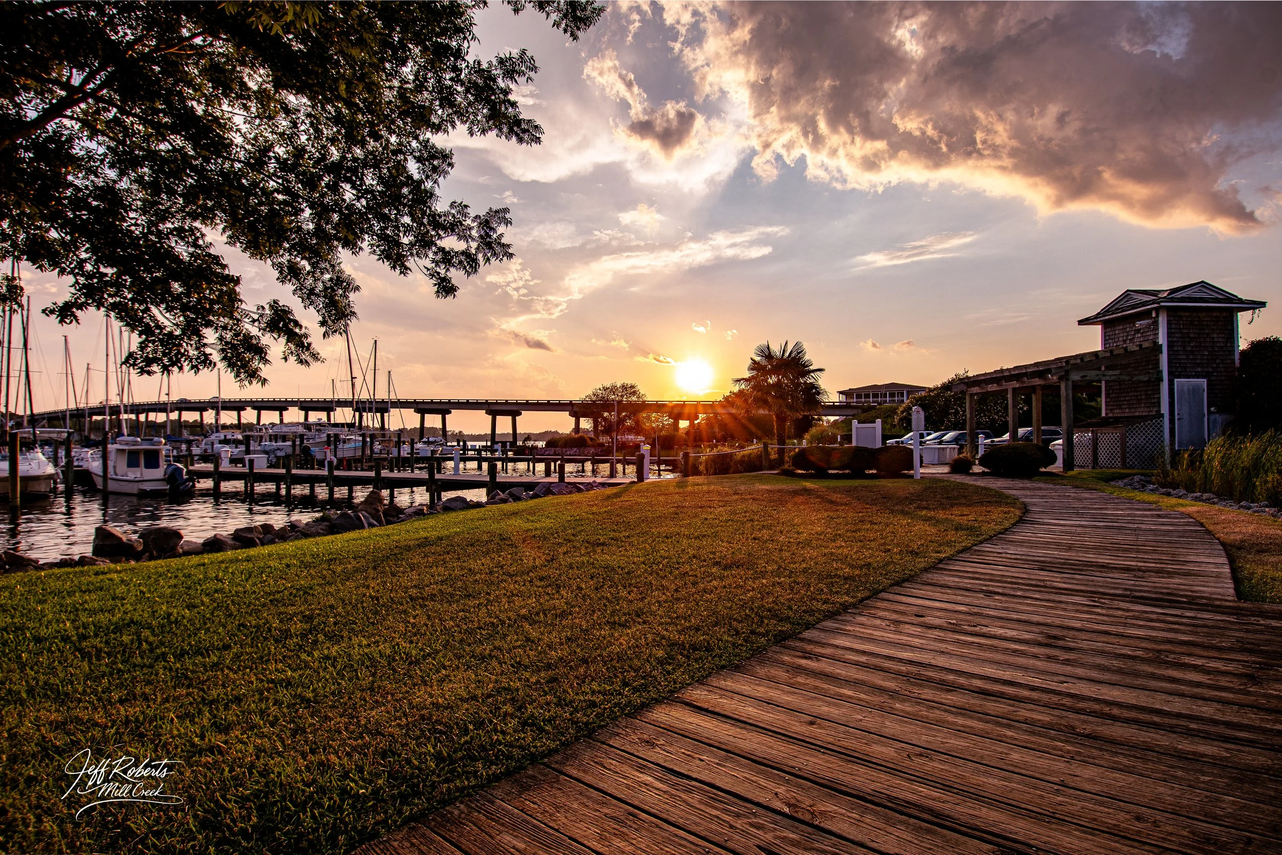 Sunset over a marina with boats docked, a wooden footpath, green grass, trees, and a sky with clouds.
