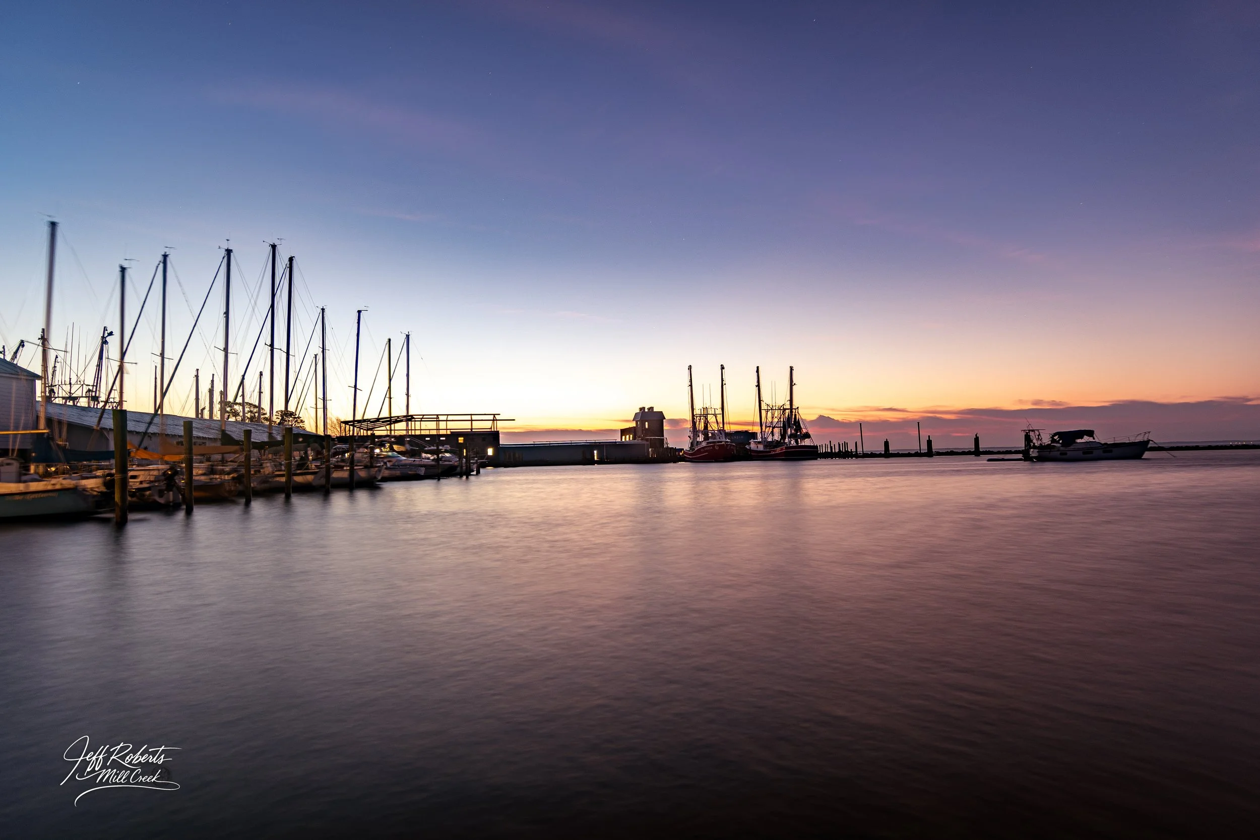 A serene marina at sunset with boats docked along the pier and calm water reflecting the colorful sky.