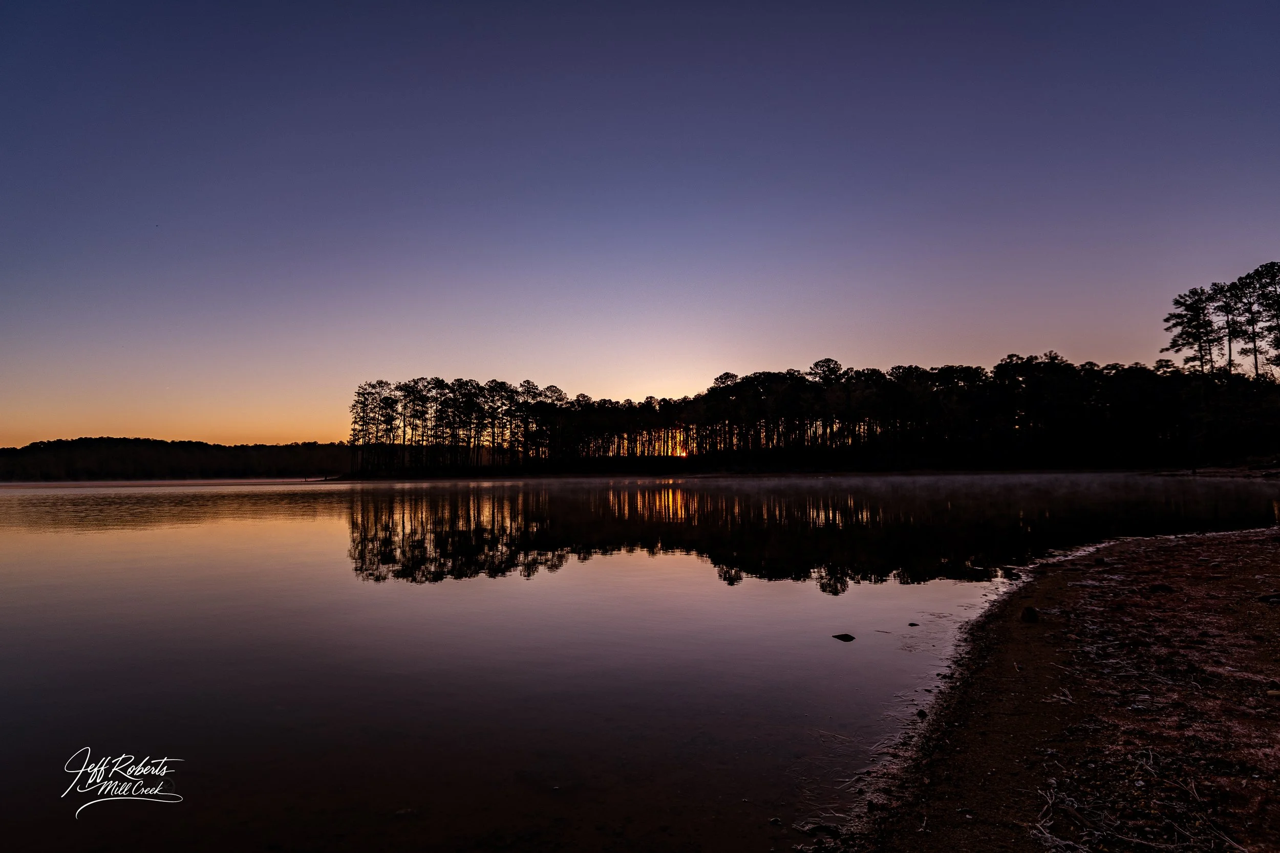 Sunset over a calm river with a silhouette of a tree-lined shoreline reflected on the water, with a clear sky transitioning from orange to purple.