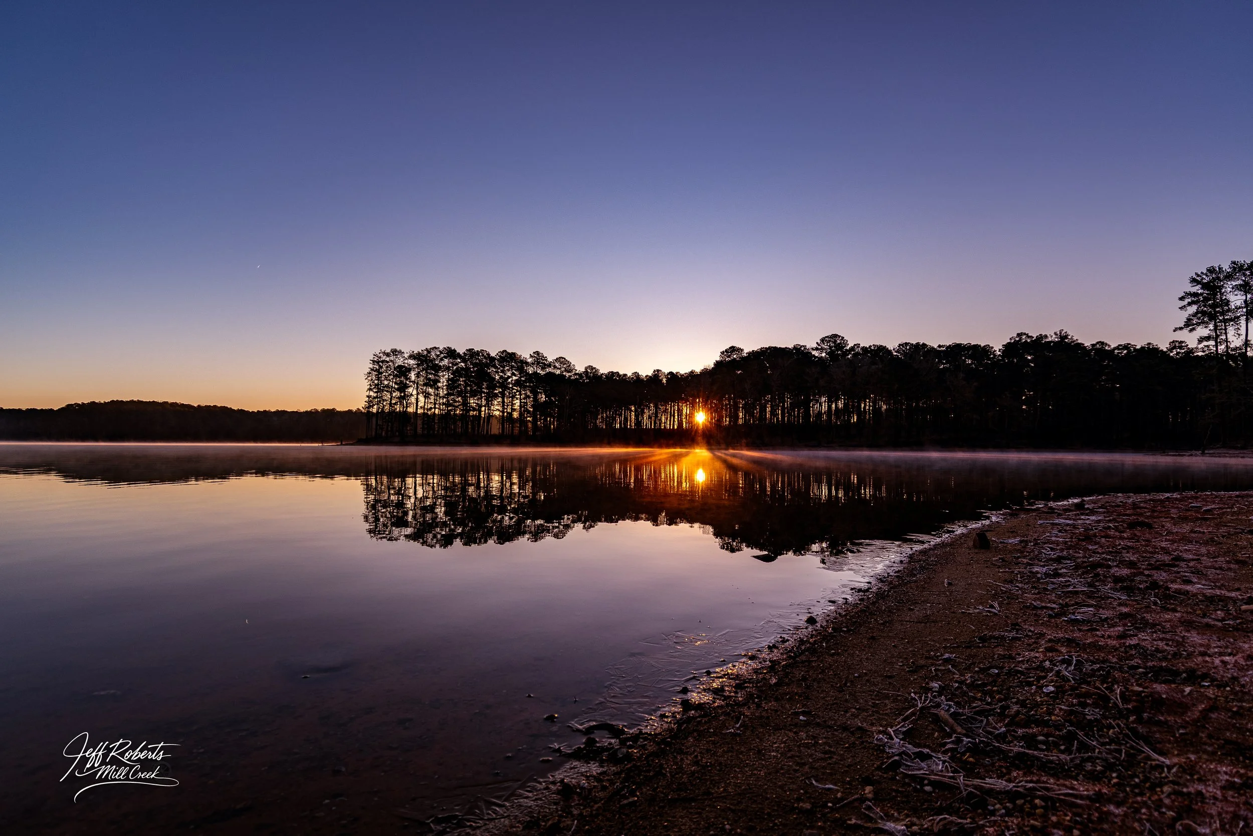 Sun setting behind a line of trees across a calm lake, creating a mirror reflection of the trees and sunset on the water, with a sandy shoreline in the foreground.