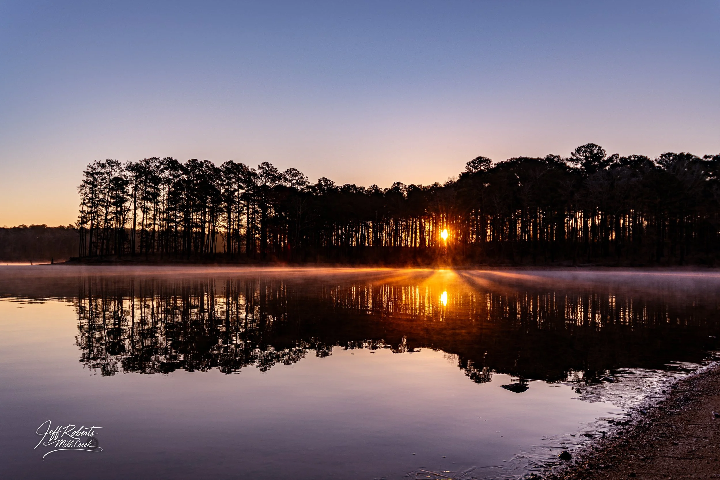 Sunrise over a calm lake with reflections of trees and mist rising from the water, signature in bottom left corner.