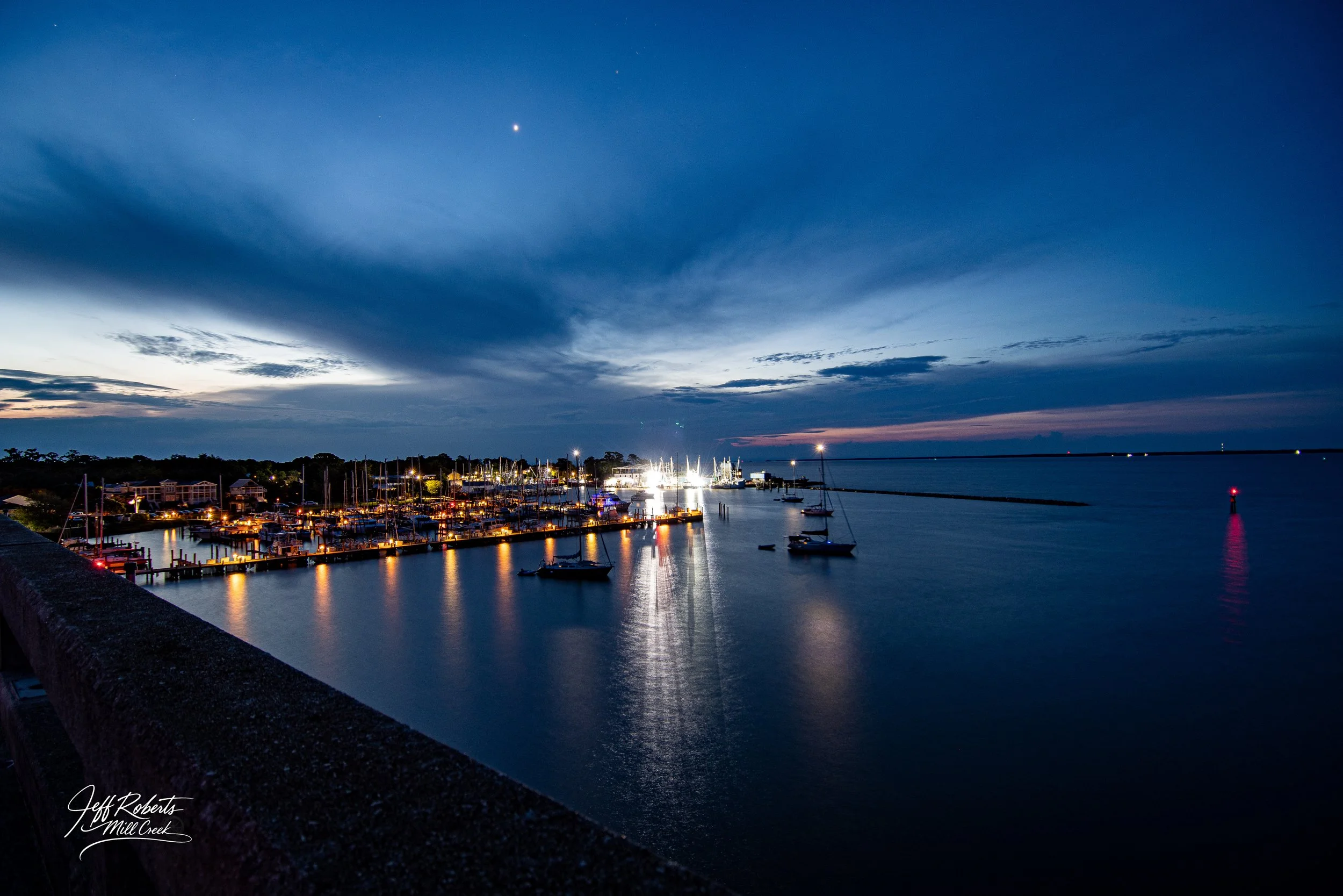 A scenic view of a marina at dusk with boats docked, illuminated by lights, under a partly cloudy sky with a visible star, reflecting on calm water.