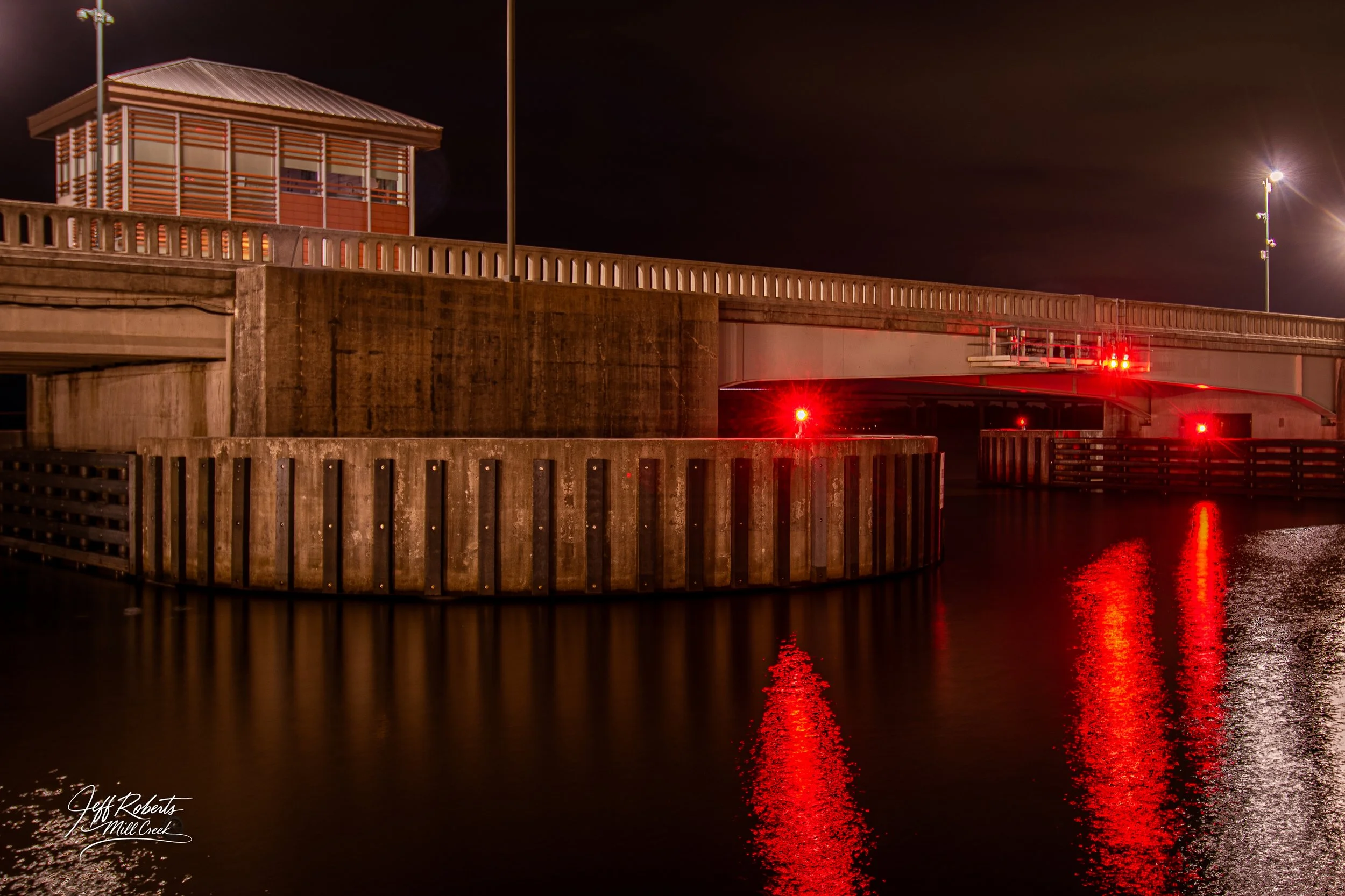 Nighttime view of a bridge over water, with red warning lights and reflections, and a small structure with a slatted roof on top.