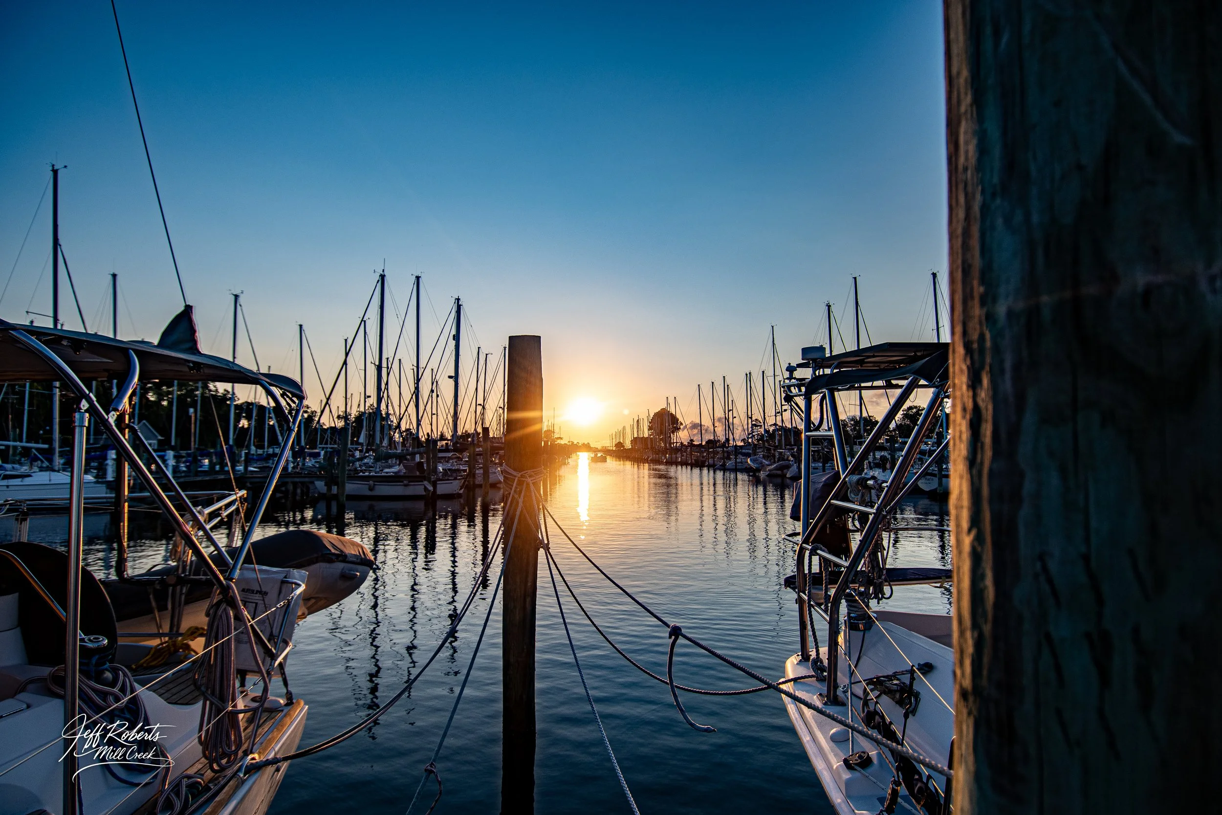 Sailboats docked at a marina during sunset with calm waters reflecting the sky and boats.
