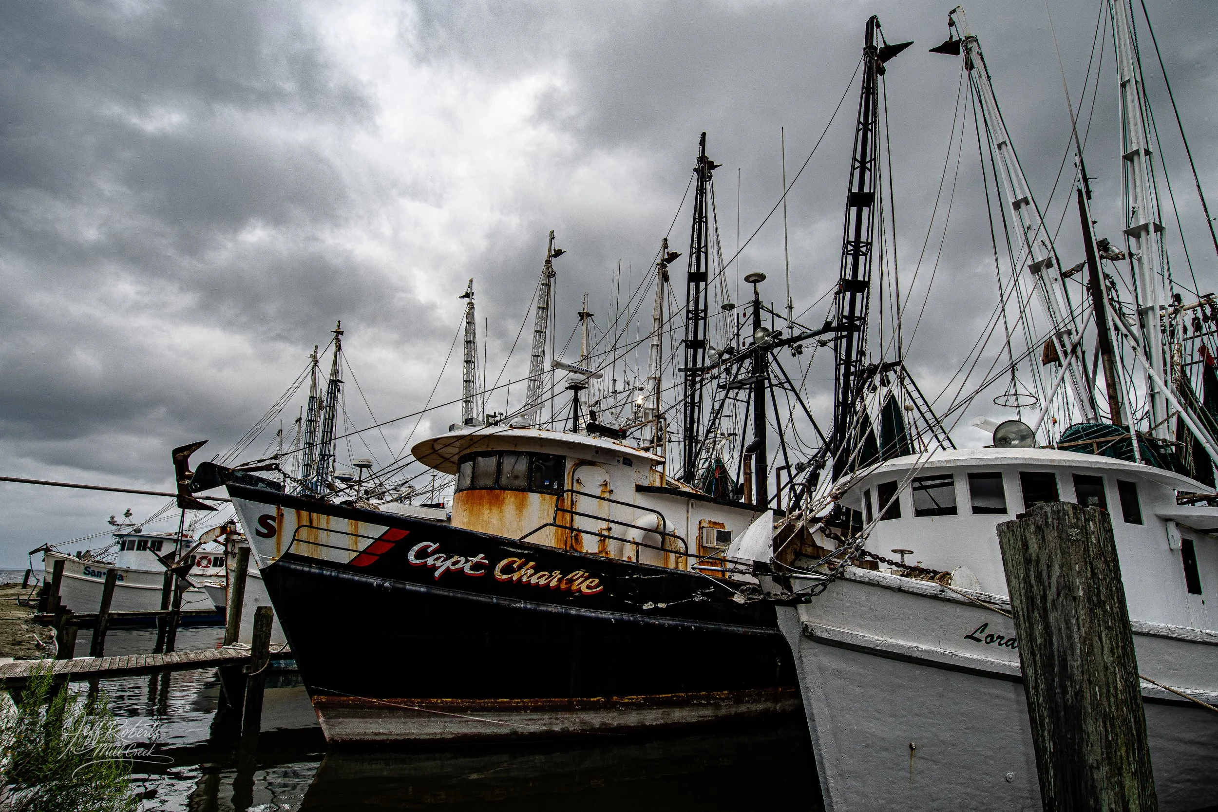 Old, rusty fishing boats docked at a marina on a cloudy day, with dark storm clouds overhead.