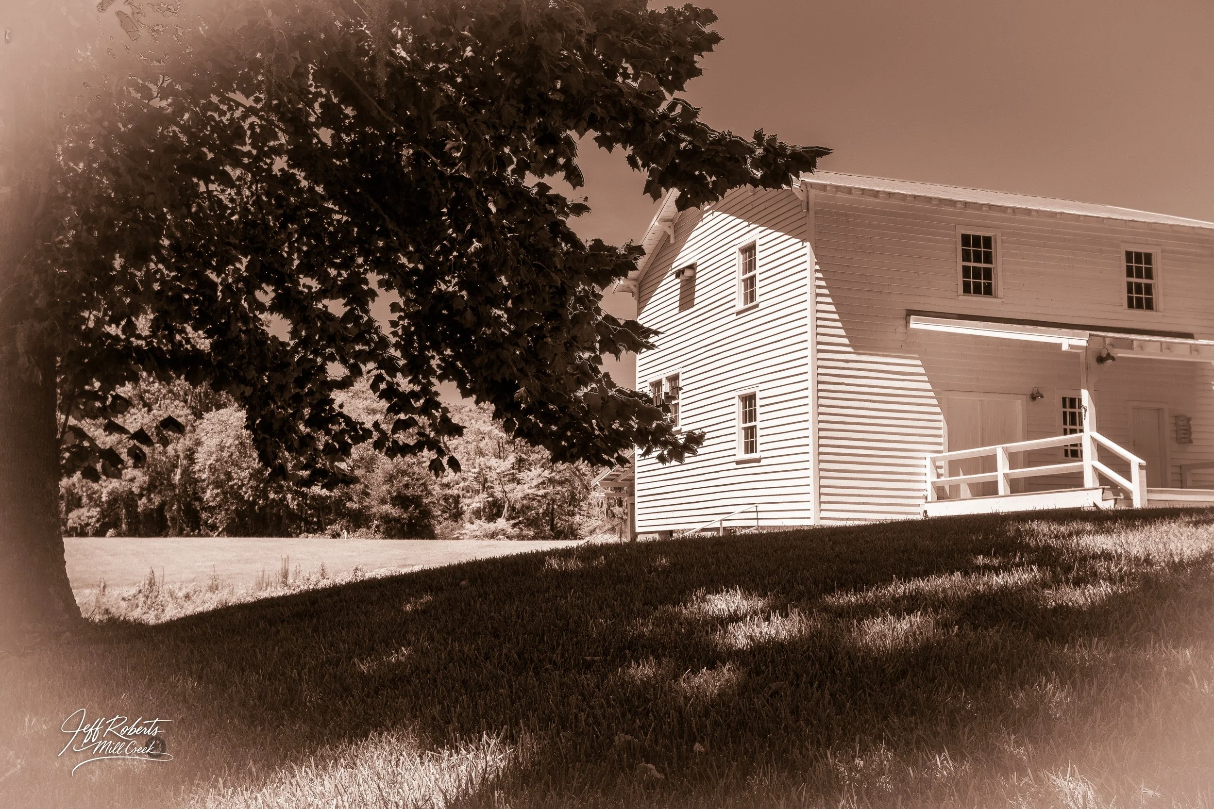 Sepia-toned photograph of a white wooden house with a porch, set on a grassy lawn, with large leafy trees in the foreground and background.