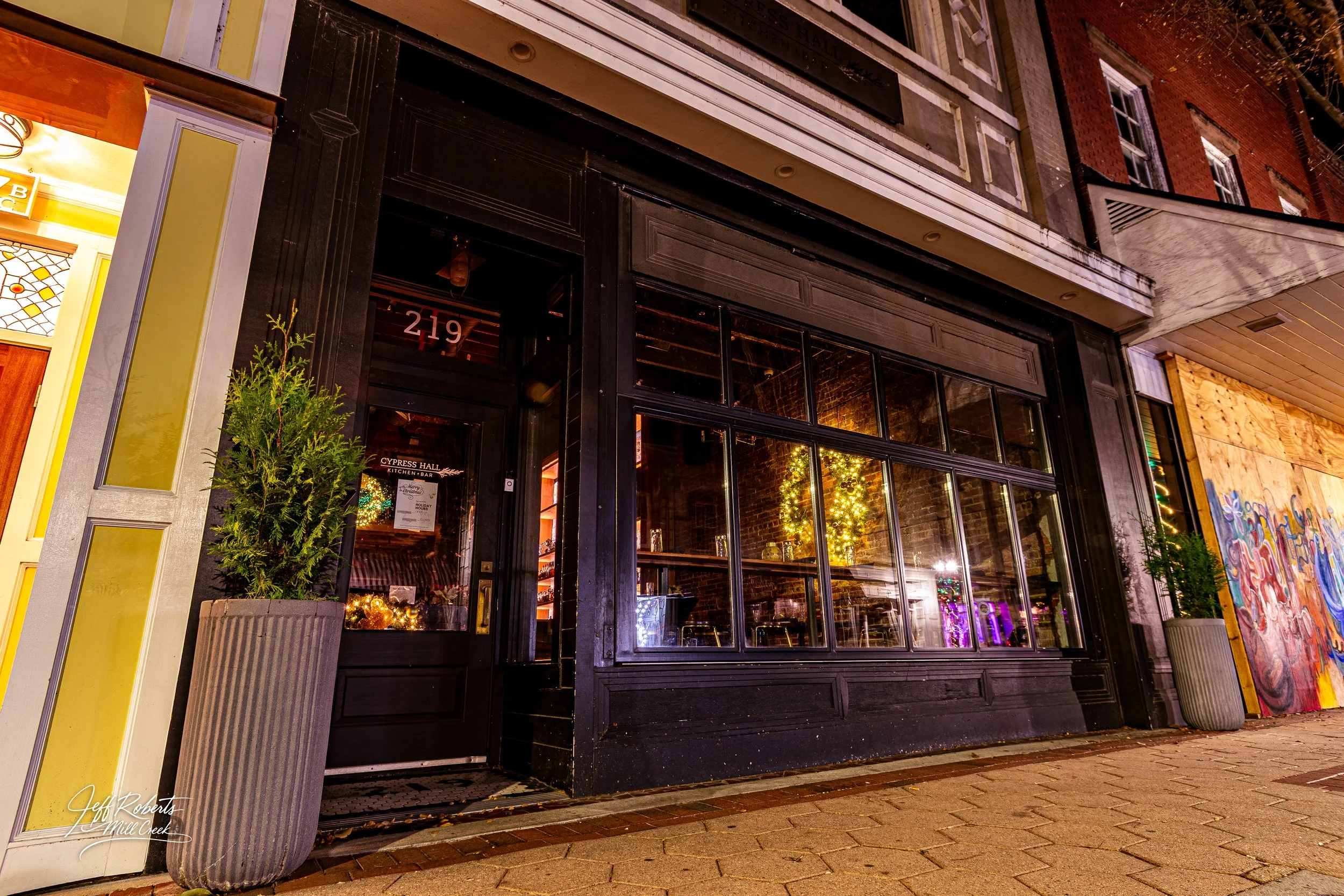 Nighttime view of the exterior of a restaurant or bar with large glass windows, black framing, and festive holiday decorations inside, including a decorated Christmas tree. Potted plants are on either side of the entrance. The street is cobblestone, 