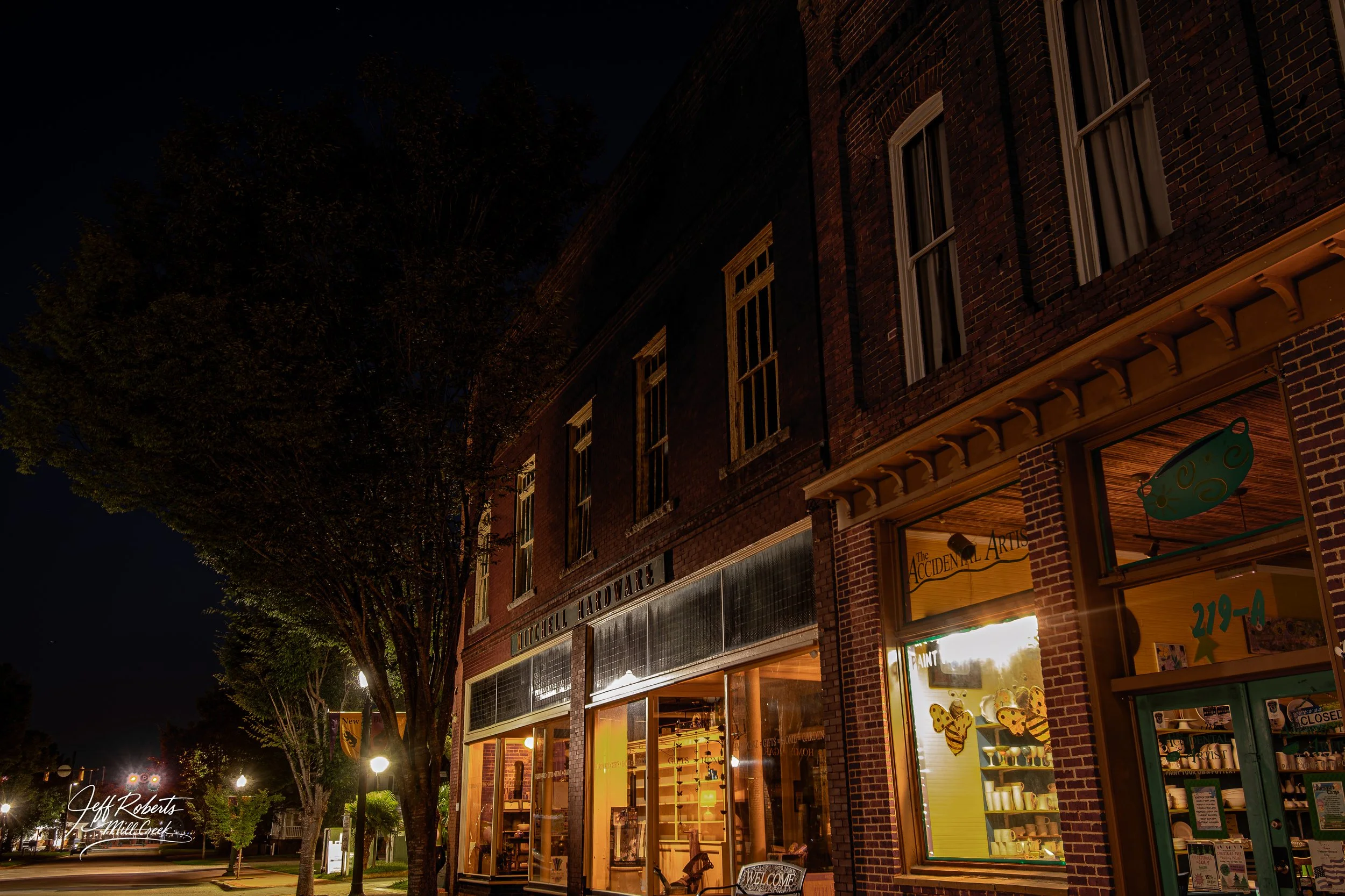 Nighttime street view of historic brick buildings, featuring a hardware store and a store named 'The Accidental Artist' with glowing interior lights, large windows, and a sign that says 'Welcome,' colorful decorations visible through the windows, and