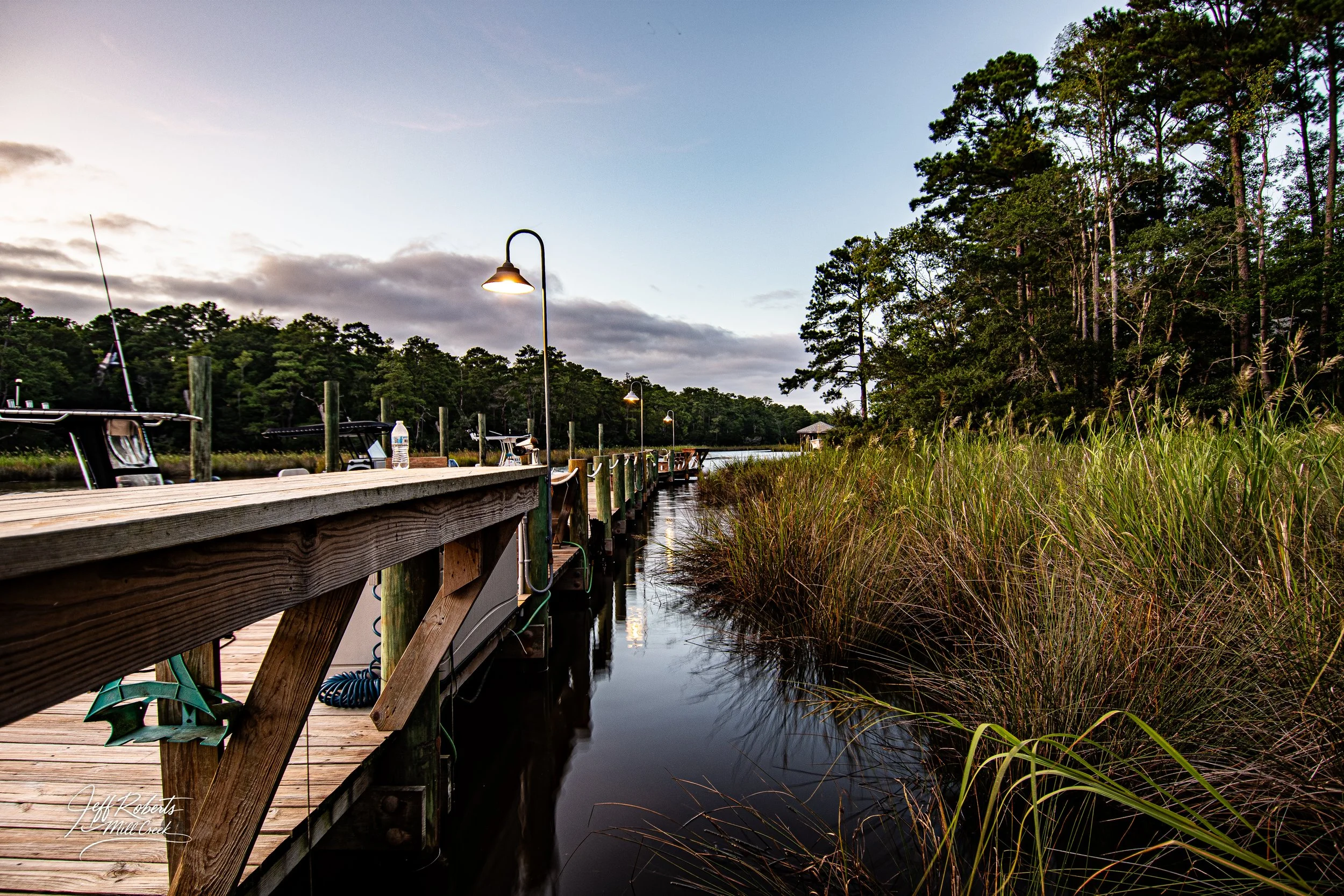 Wooden dock extending into a calm waterway, with boats moored on one side. Tall grass and trees border the water, under a mostly cloudy sky with some light from lamps along the dock.