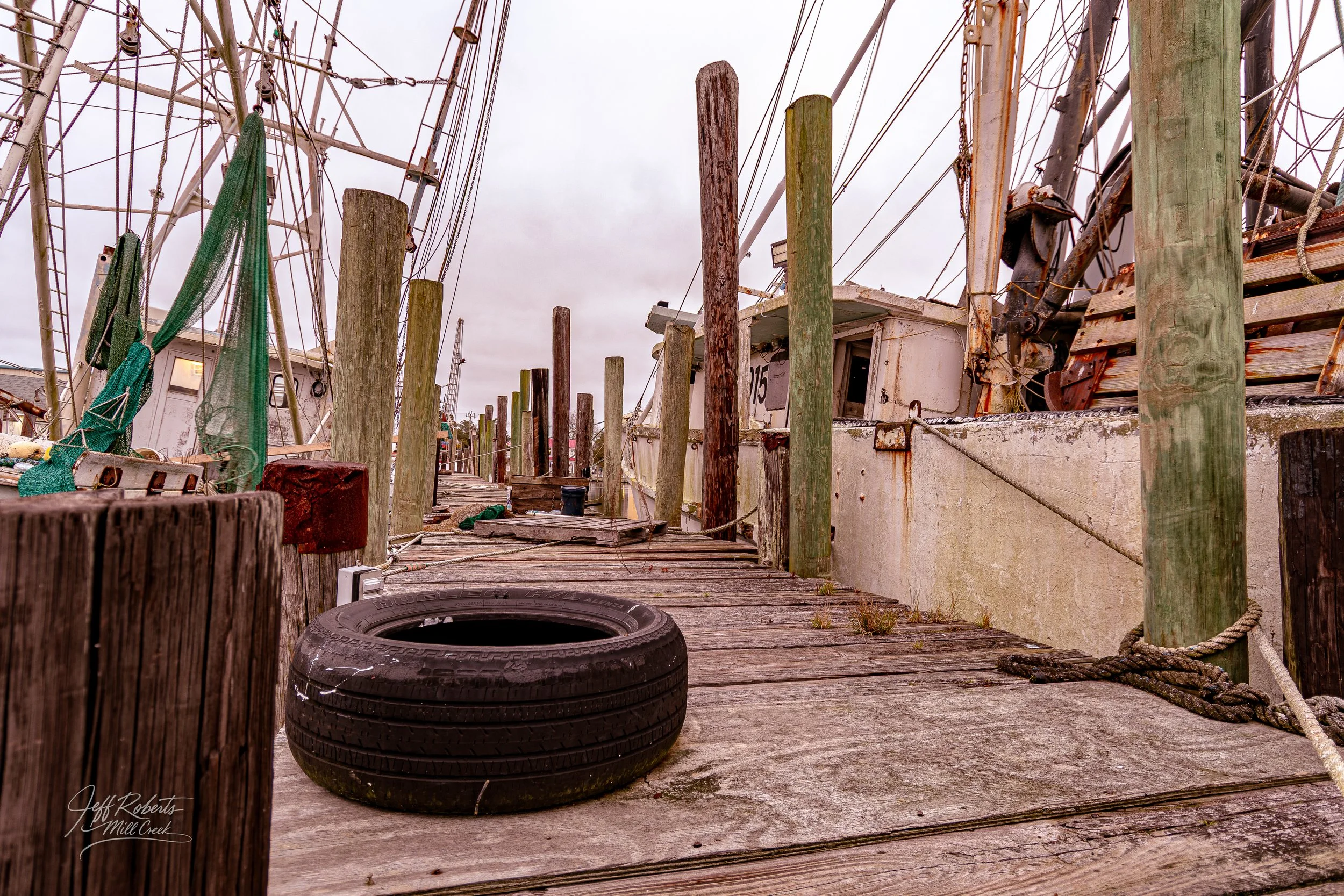 View of a weathered dock with a torn tire used as a bumper, old wooden posts and a large boat with rust and peeling paint moored alongside, overcast sky in the background.