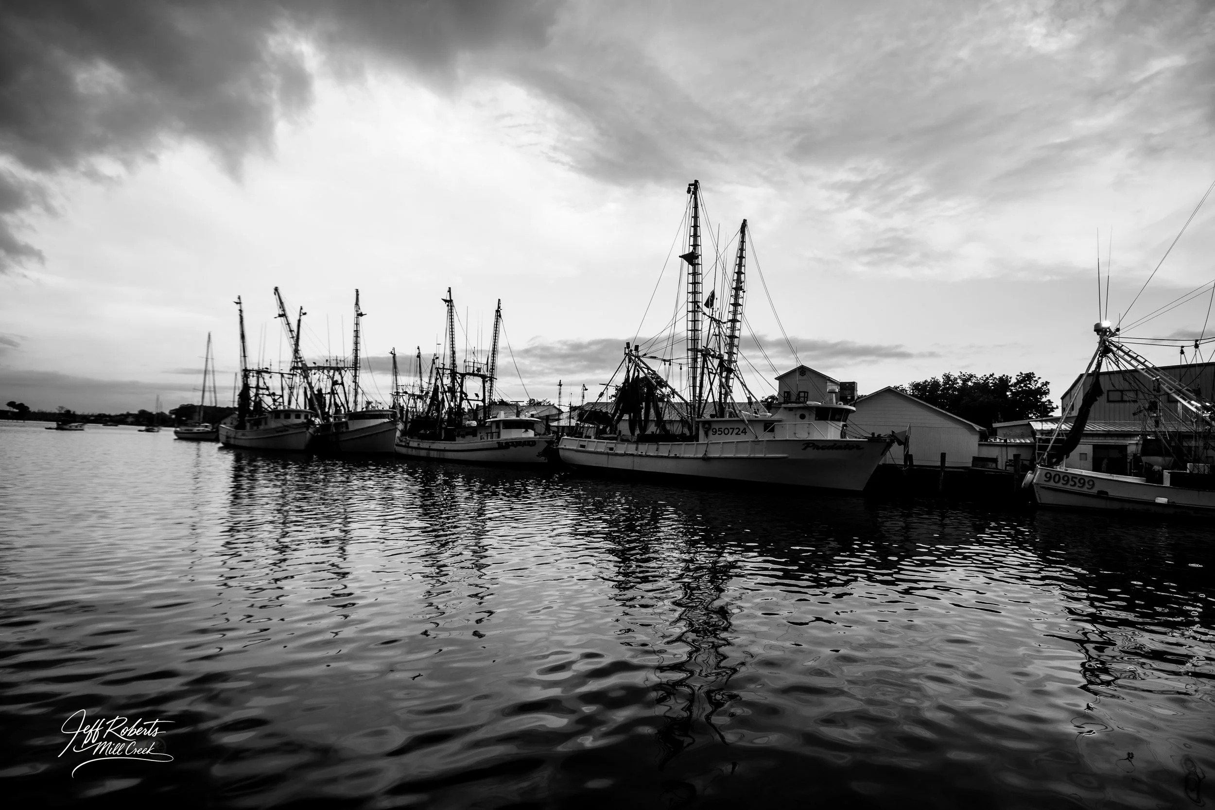 A black and white photo of several boats docked at a marina with reflections on the water and cloudy sky above.