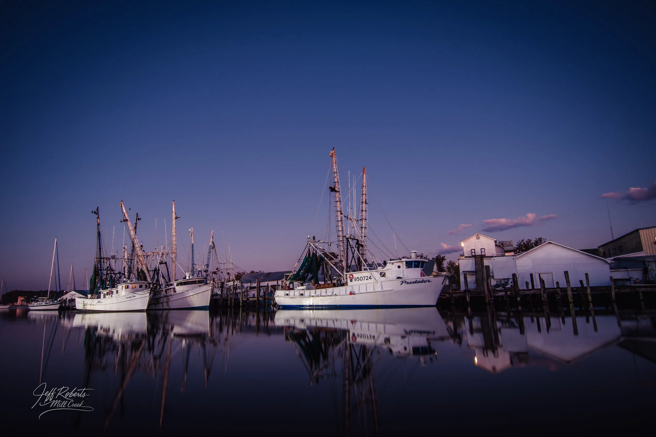 Nighttime scene of sailboats docked at a harbor, with their reflections visible in calm water, and a few clouds in the sky.