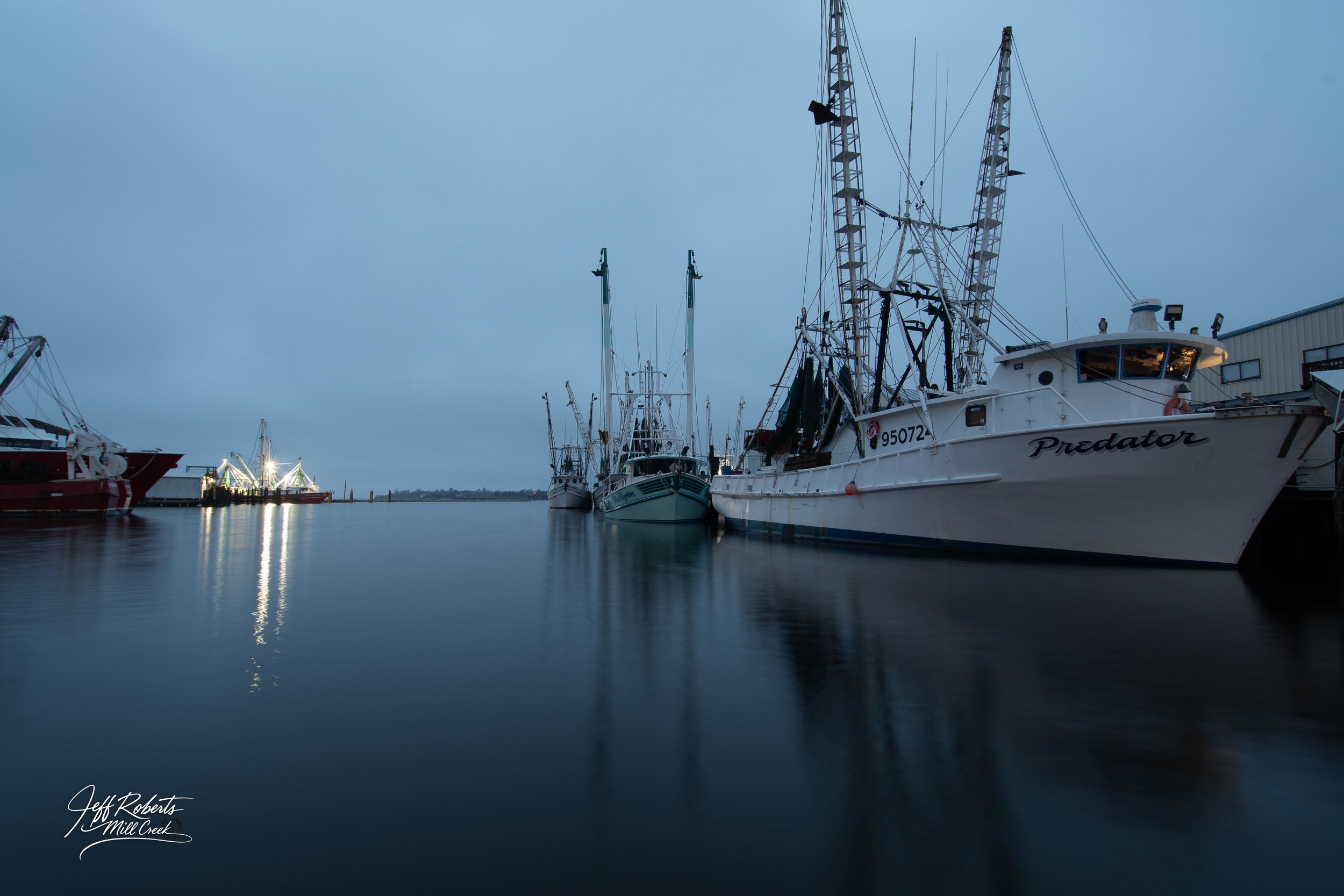 A marina with several fishing boats docked, calm water reflecting the ships, and a cloudy sky in the background.