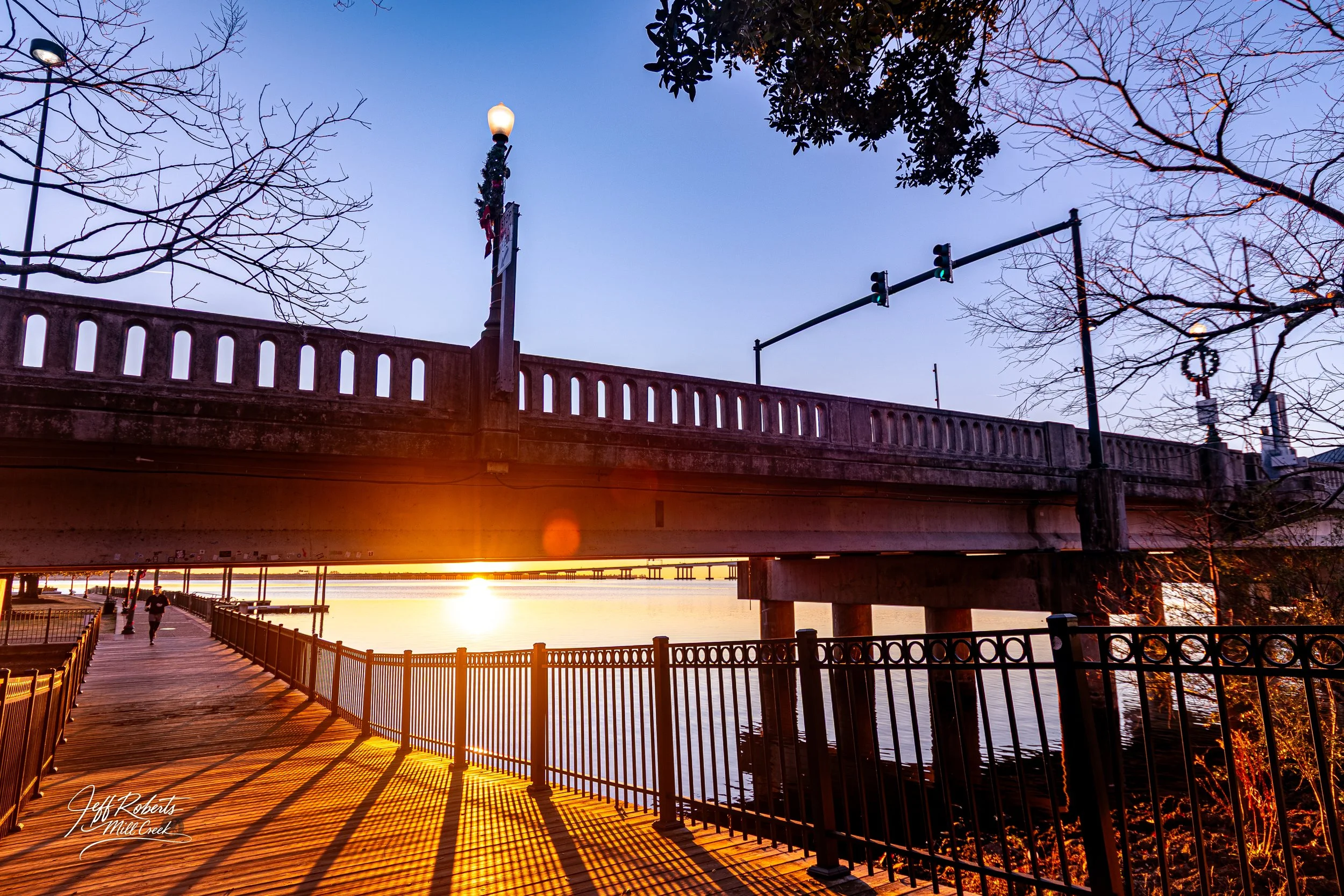 A sunset view beneath a bridge over a river with a boardwalk, railing, and a person jogging along the path.