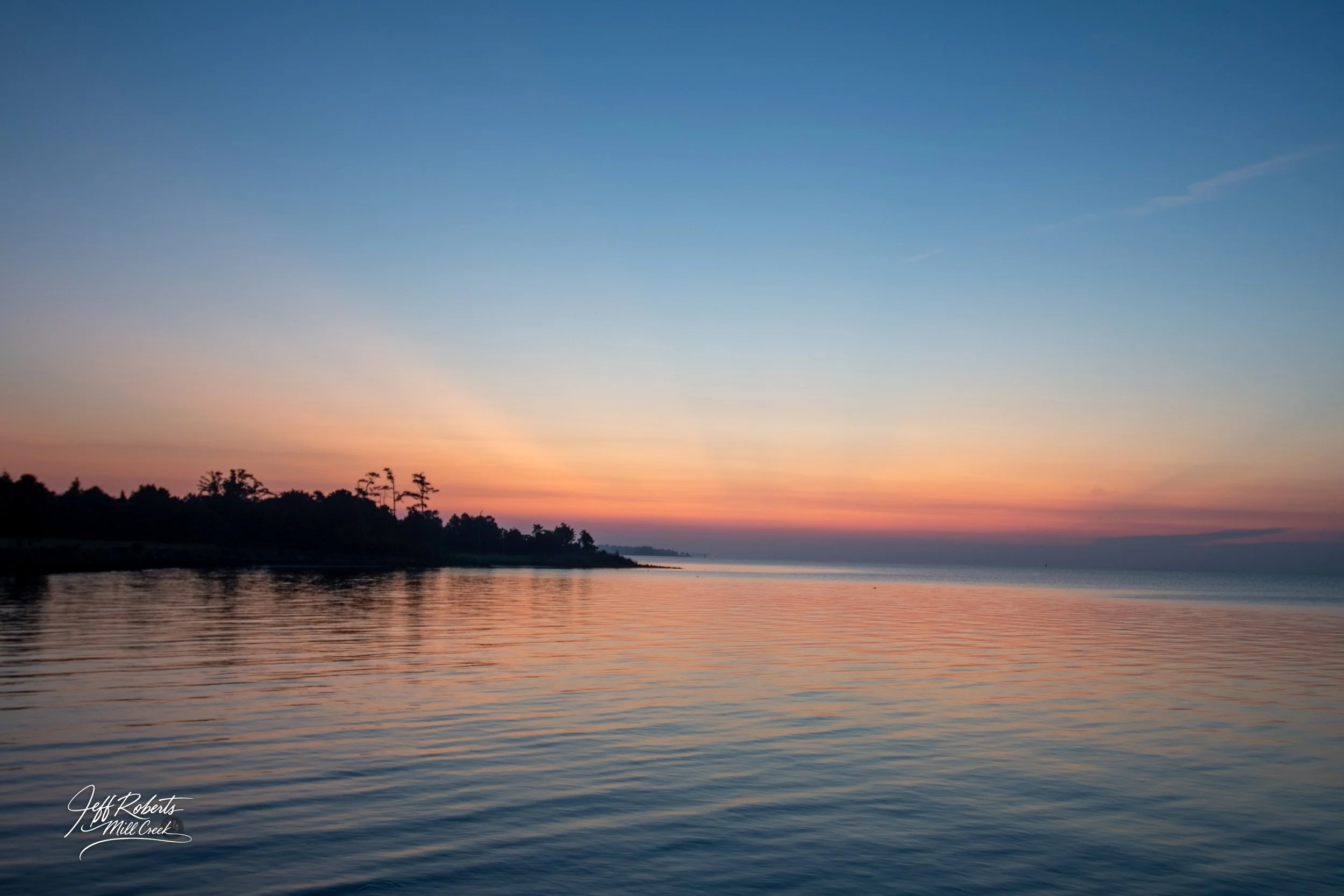 Sunset over a calm body of water with trees along the shoreline and a colorful sky transitioning from orange to blue.