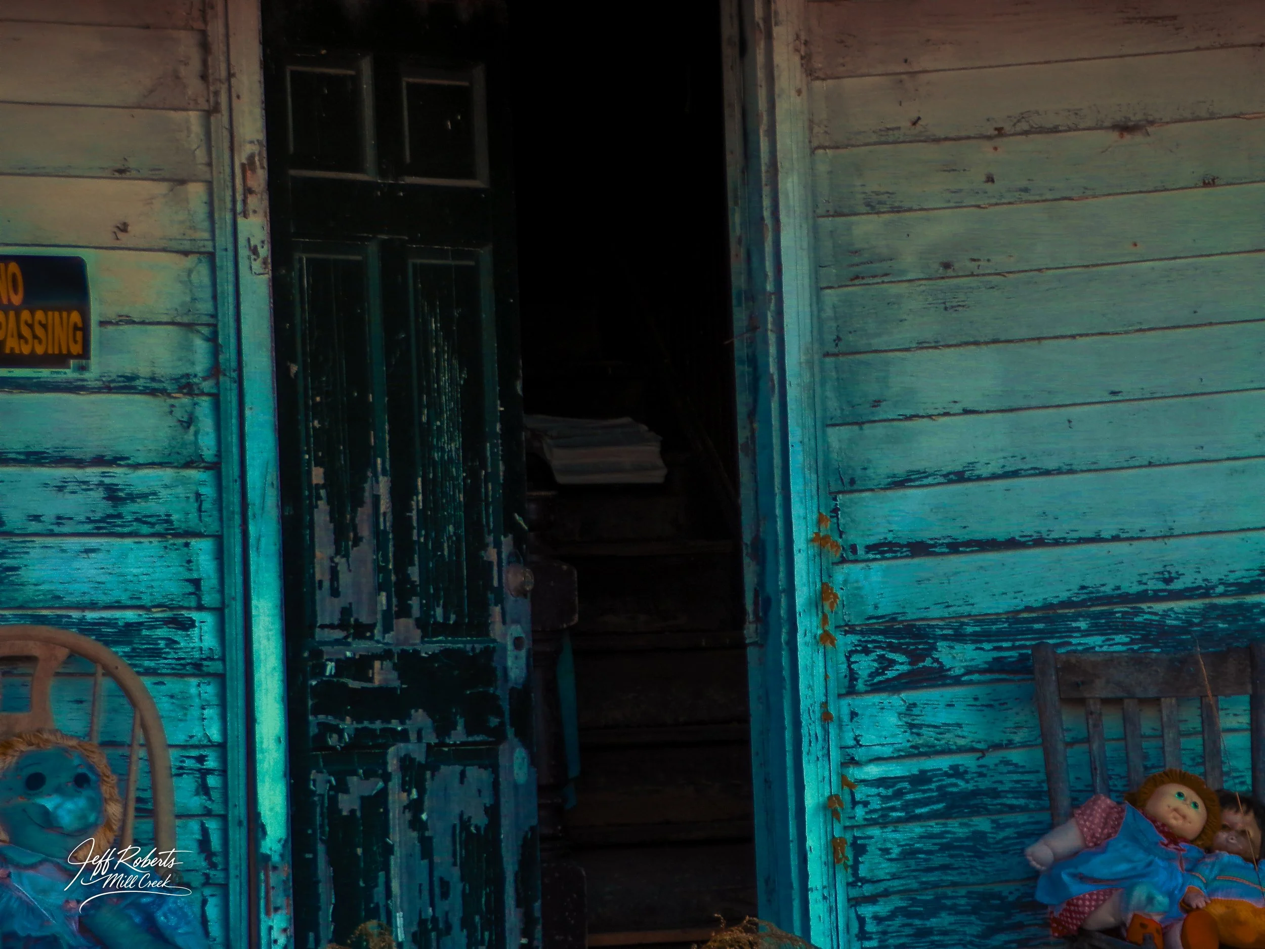 An open door leading into a dark interior of an old, weathered blue wooden house with peeling paint. To the left and right of the door are vintage dolls sitting on chairs, with the house's exterior showing signs of age and neglect.