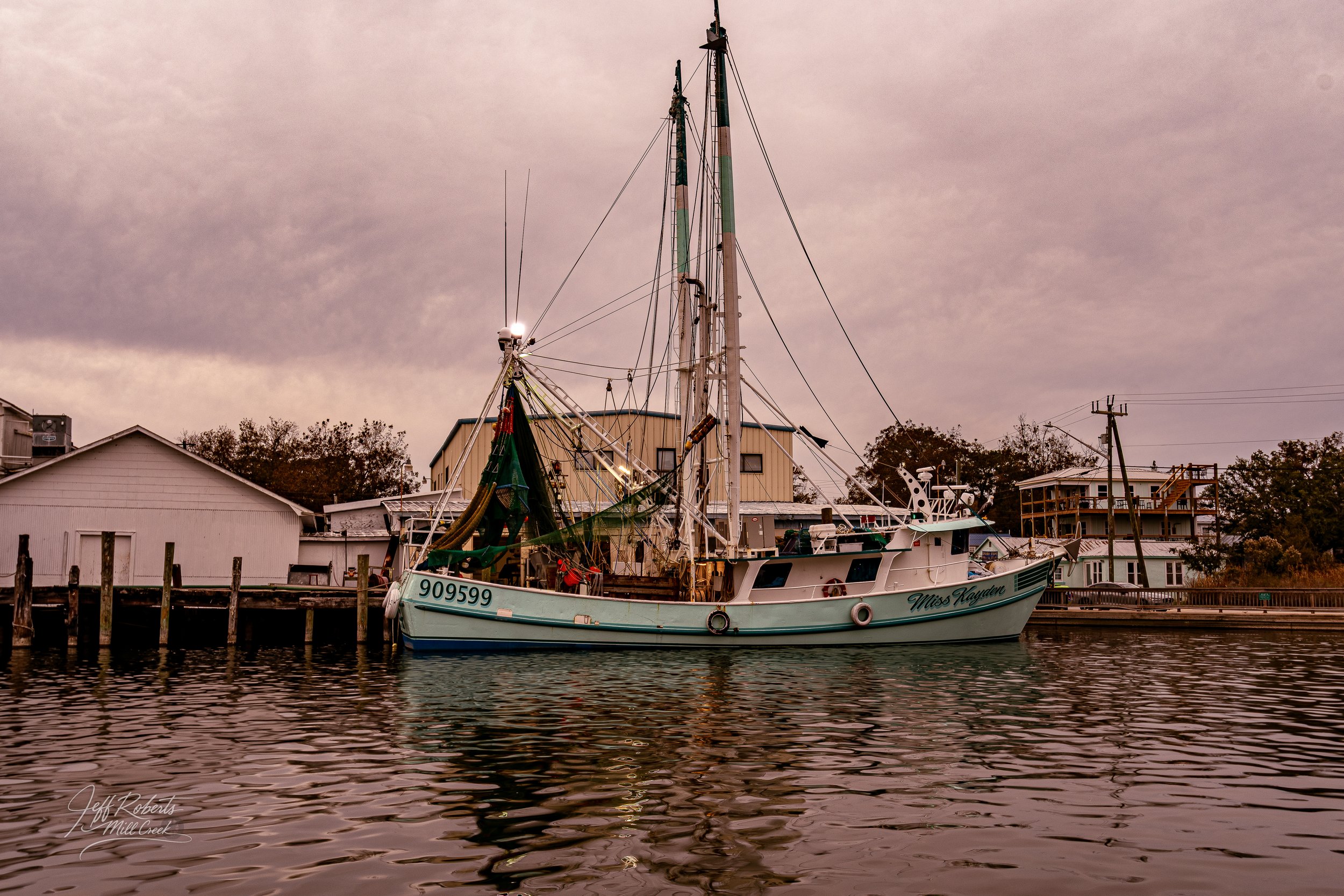 A fishing boat named Miss Kayden docked along a waterfront with residential houses in the background, under a cloudy sky.