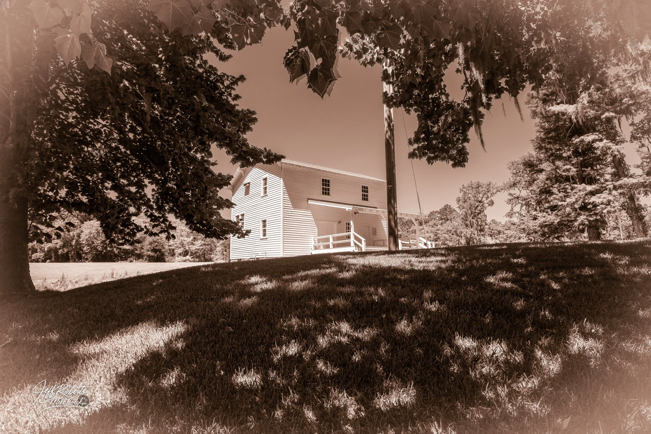 A white wooden barn on a grassy hill surrounded by trees, with power lines and a utility pole in the foreground and a clear sky.