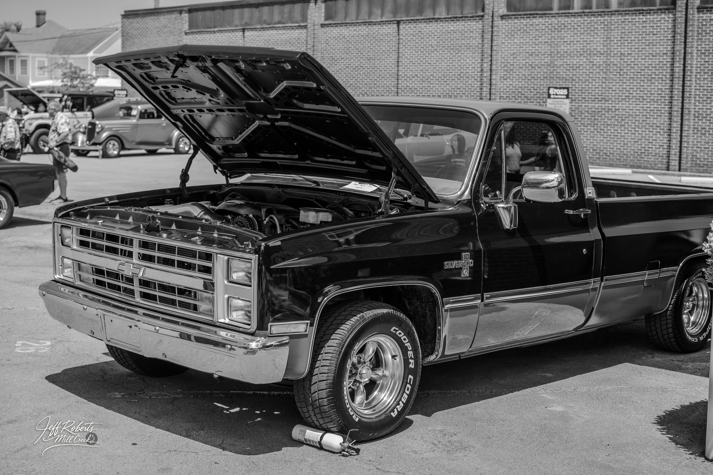 Black vintage Chevrolet Silverado pickup truck with open hood, parked at an outdoor car show, with other classic cars and people in the background.