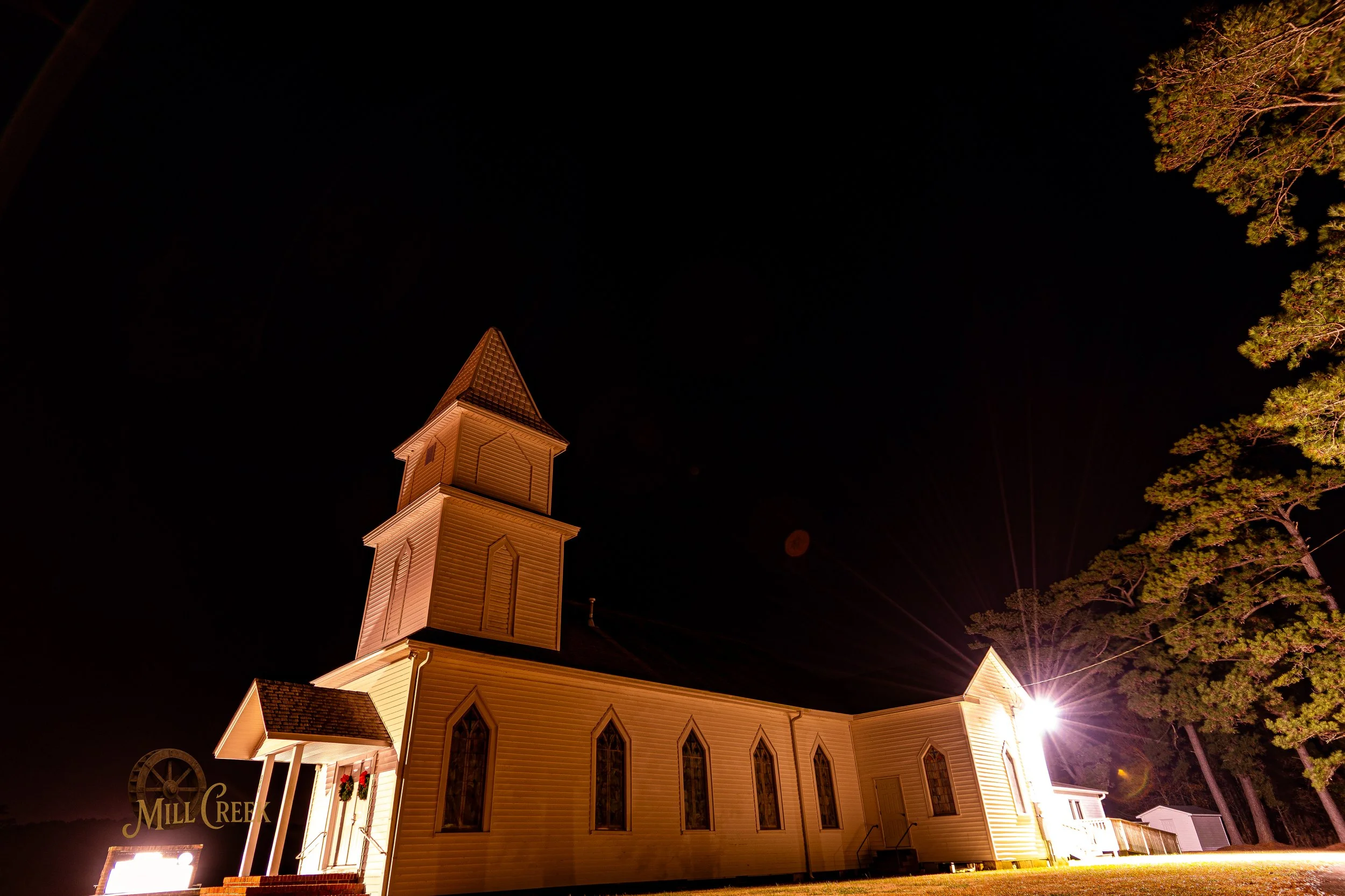 Nighttime view of a wooden church with tall steeple, illuminated by bright exterior light, with several narrow stained glass windows and a windmill structure nearby, and tall trees in the background.