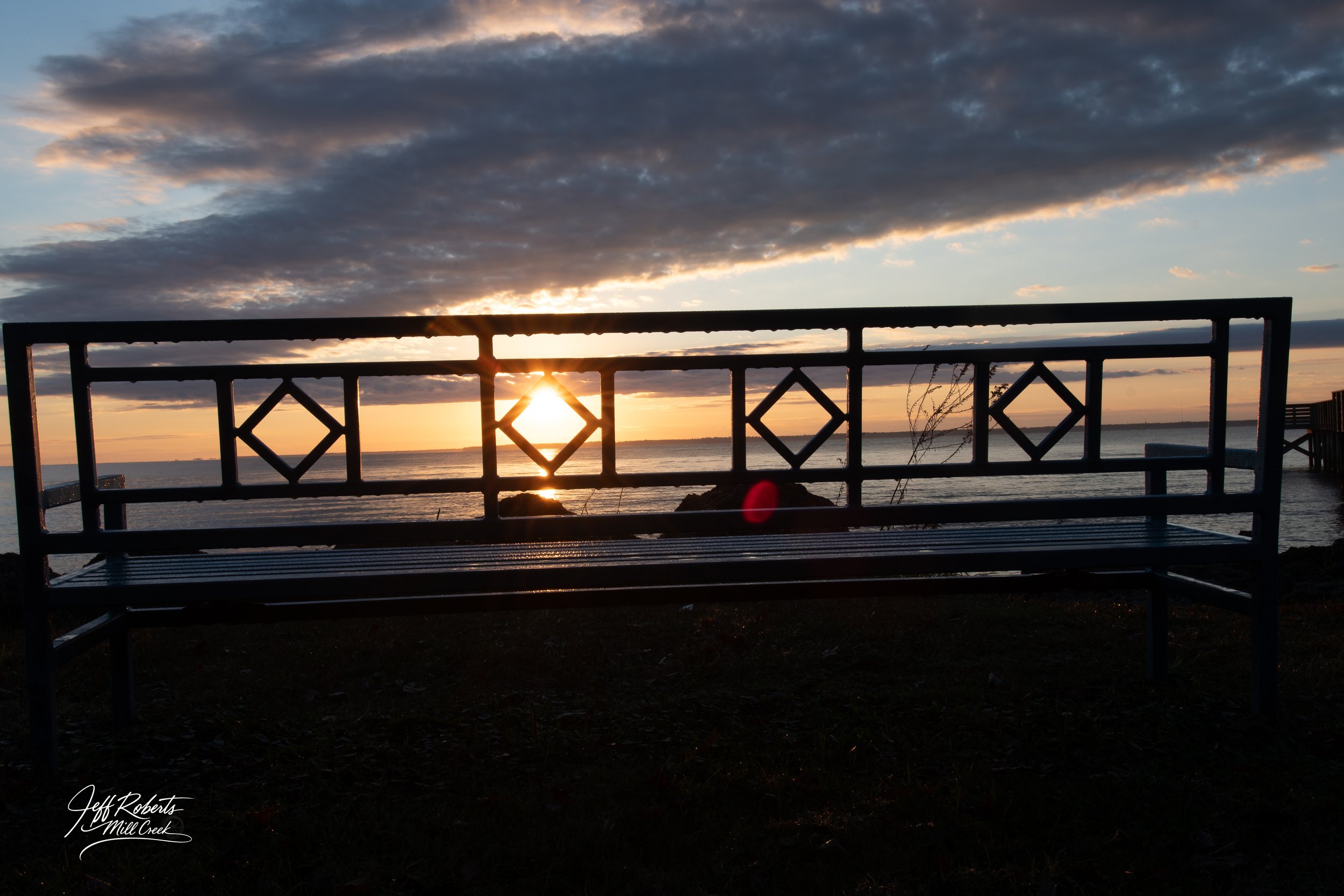 Silhouette of a park bench facing the water at sunset with clouds in the sky.
