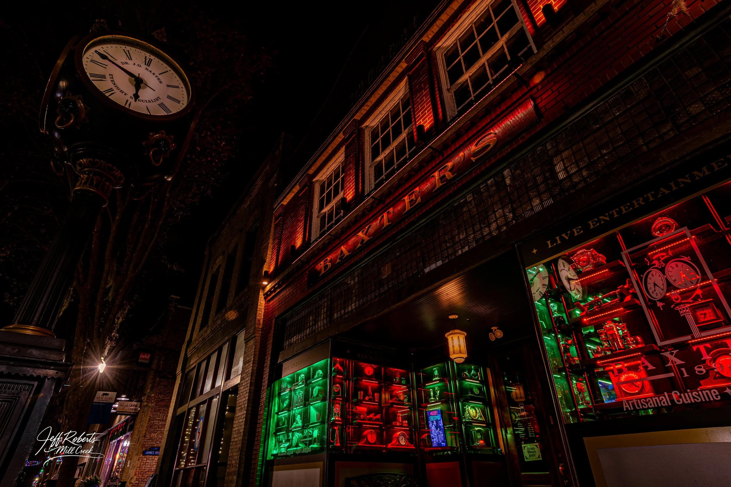 Nighttime exterior view of a brick building with neon red and green signs and displays, including clocks and various decorative items, under a dark sky with streetlights visible.