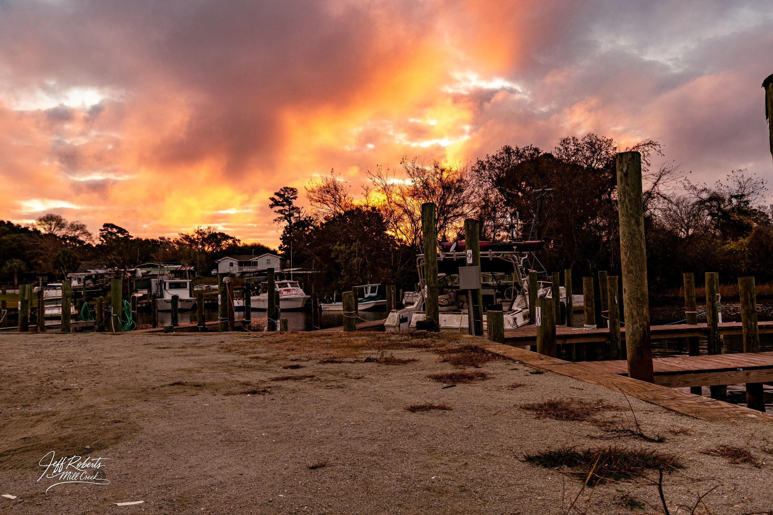 Sunset over a marina with boats docked at wooden piers and trees in the background.