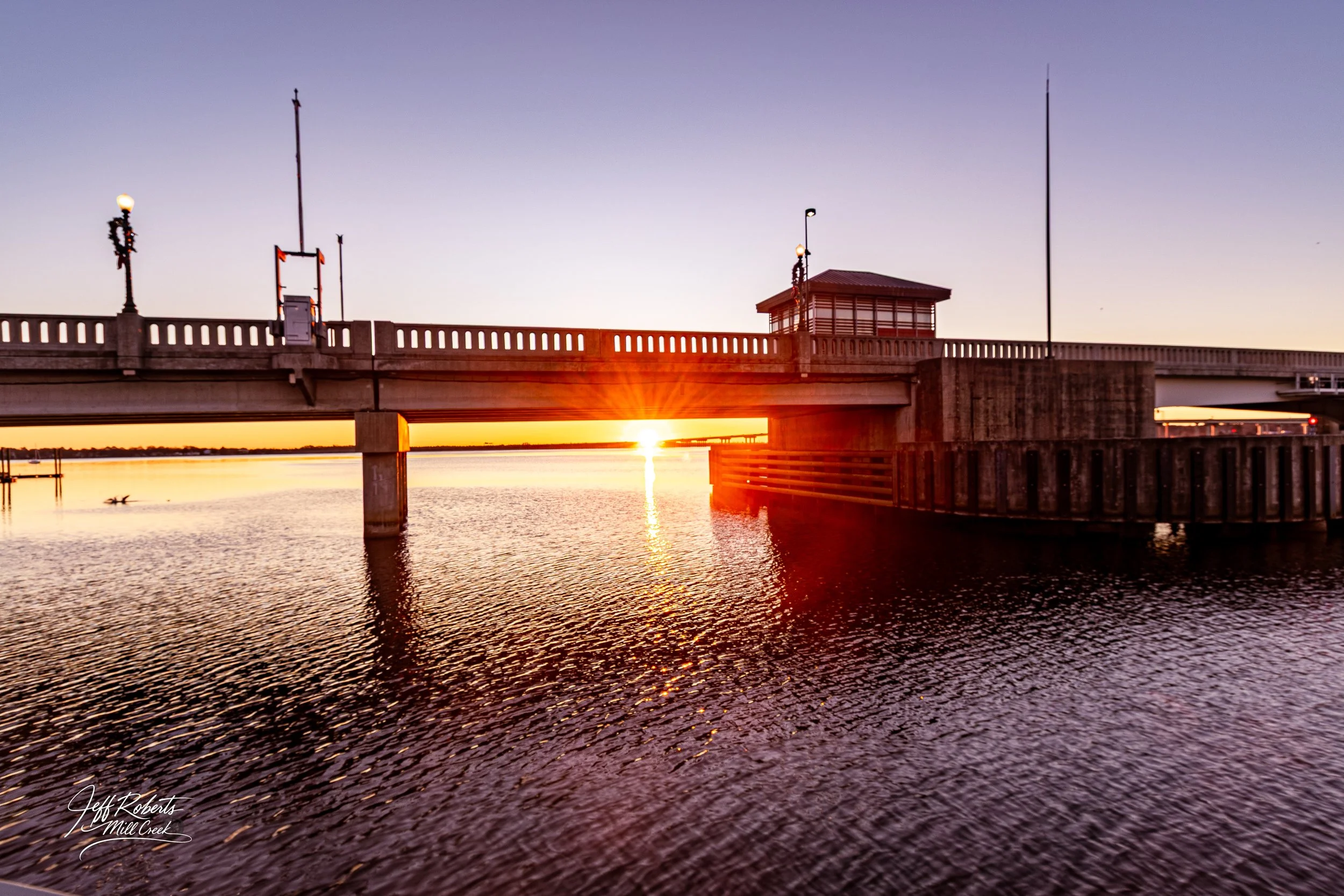 Sunset over a waterway with a bridge, small structure, and reflections in the water.