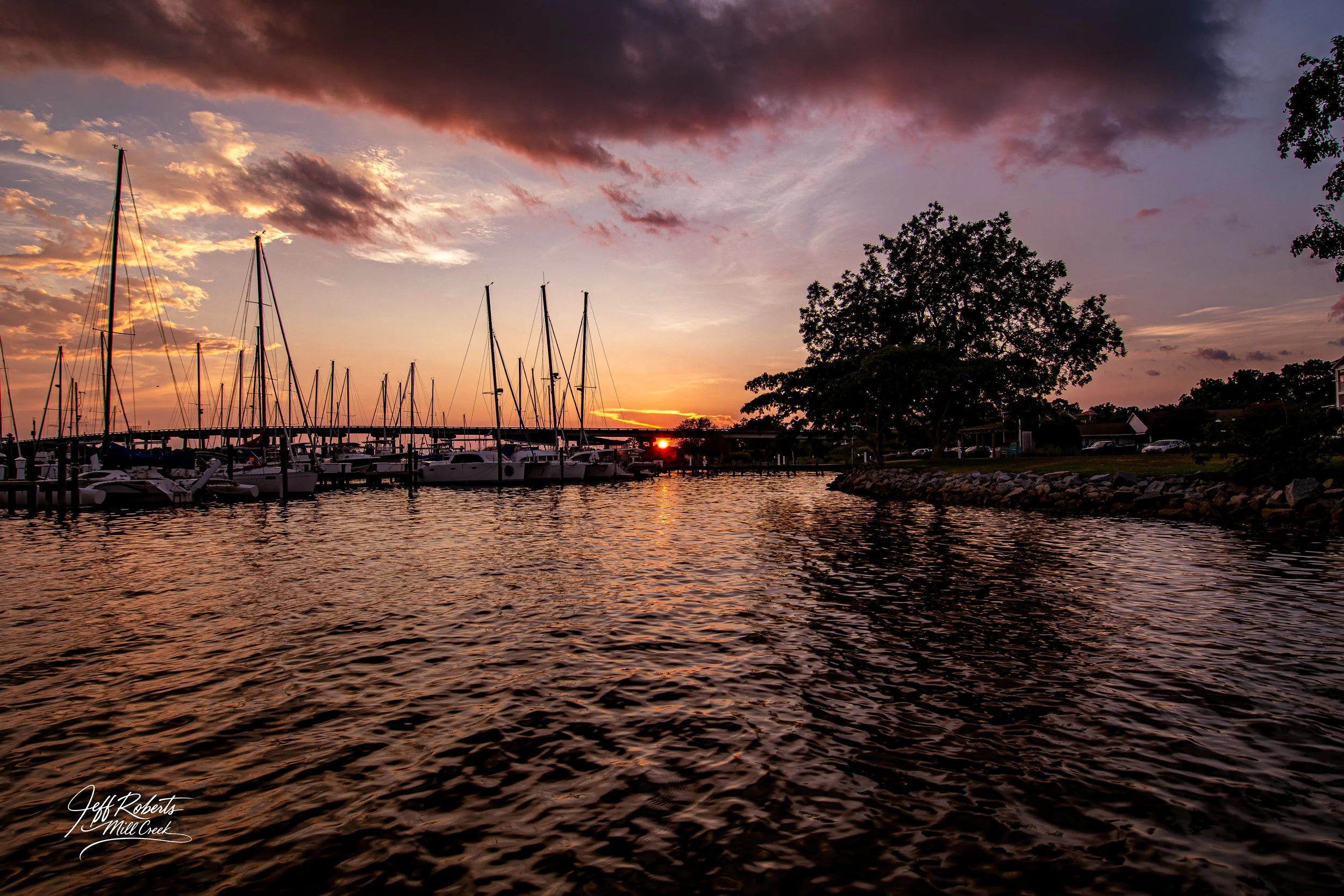 Sailboats docked at a marina during sunset with a sky filled with clouds and trees along the shoreline.