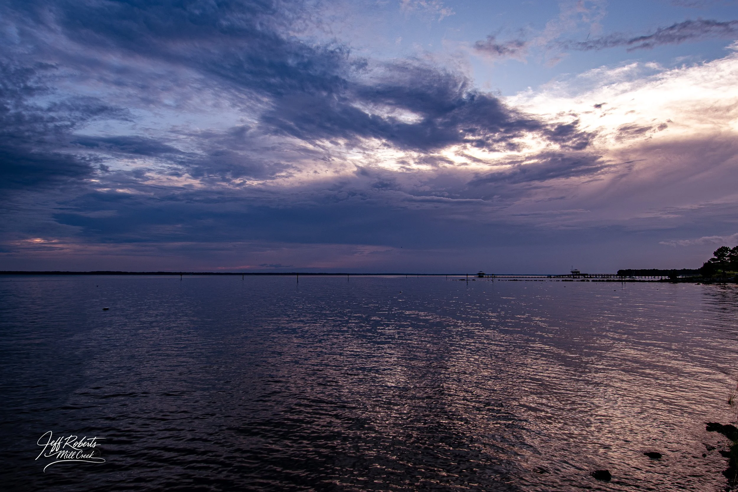 A tranquil body of water at dusk with a cloudy sky overhead and a distant shoreline with structures and trees.