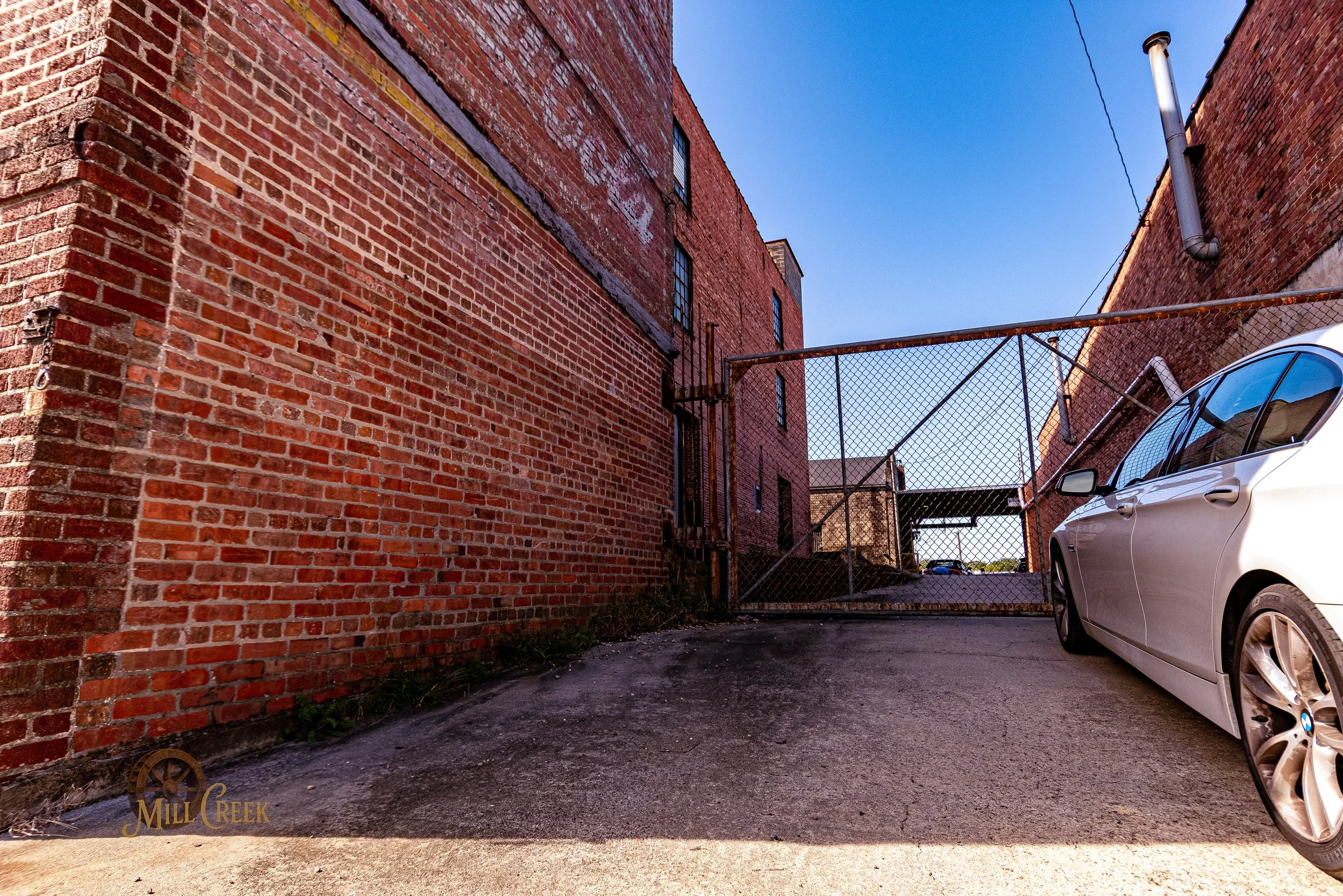 Back alley scene with brick buildings, a chain-link fence gate, a white car parked on the right, and a clear blue sky.
