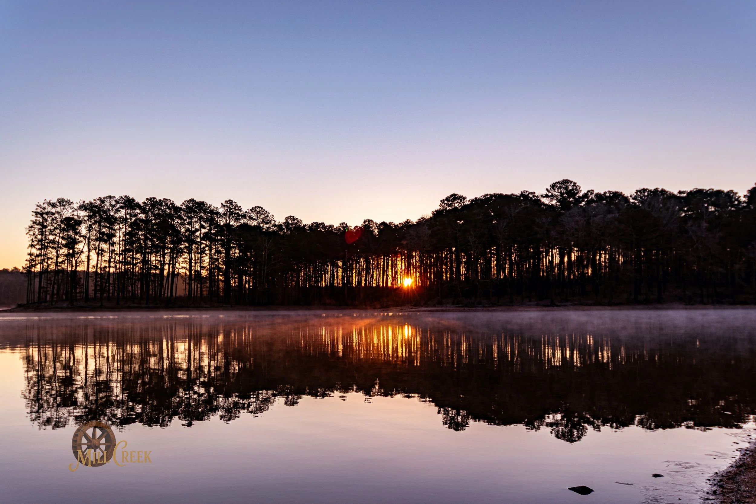 Sunset over a forested lake with mist rising from the water, reflected trees, and a clear sky.